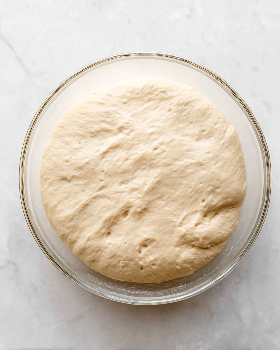 The image shows a clear glass bowl filled with a single large layer of risen dough. The dough is pale beige with a smooth and slightly wrinkled texture, showing some small air bubbles on its surface. The bowl is placed on a white marbled texture surface, and the dough fills most of the bowl's round bottom, gently rising upwards towards the edges. The photo taken with an iphone --ar 4:5 --v 7