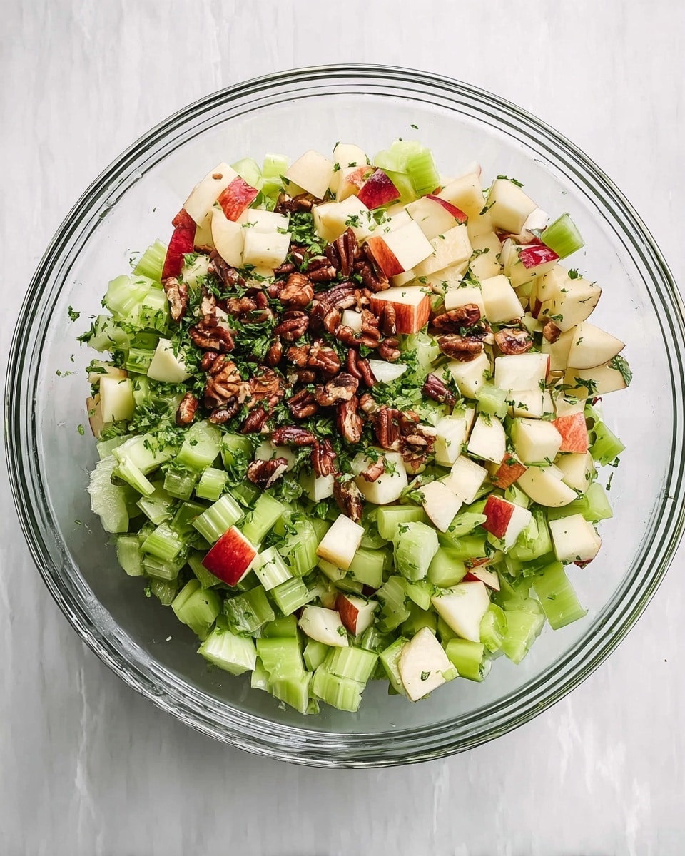 A clear glass bowl filled with a fresh salad showing about three layers of ingredients: the bottom layer has light green chopped celery pieces with a crisp texture, the middle layer includes small diced apple chunks with red skin and white inside, and the top layer is scattered with dark brown chopped nuts and green chopped herbs sprinkled evenly. The bowl is placed on a white marbled surface, enhancing the fresh colors of the salad. Photo taken with an iphone --ar 4:5 --v 7