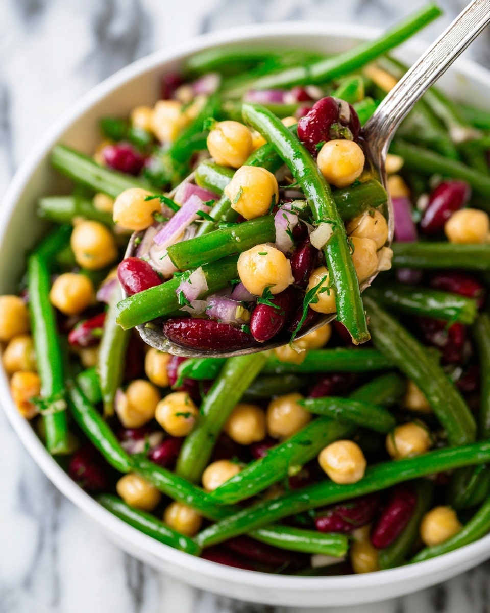 A white bowl full of three types of beans: bright green cut green beans, round yellow chickpeas, and red kidney beans, all mixed together with small pieces of thinly sliced purple onion and sprinkled with finely chopped green herbs, with a close-up silver spoon lifting a mix of the beans and onions from the bowl, set on a white marbled surface photo taken with an iphone --ar 4:5 --v 7
