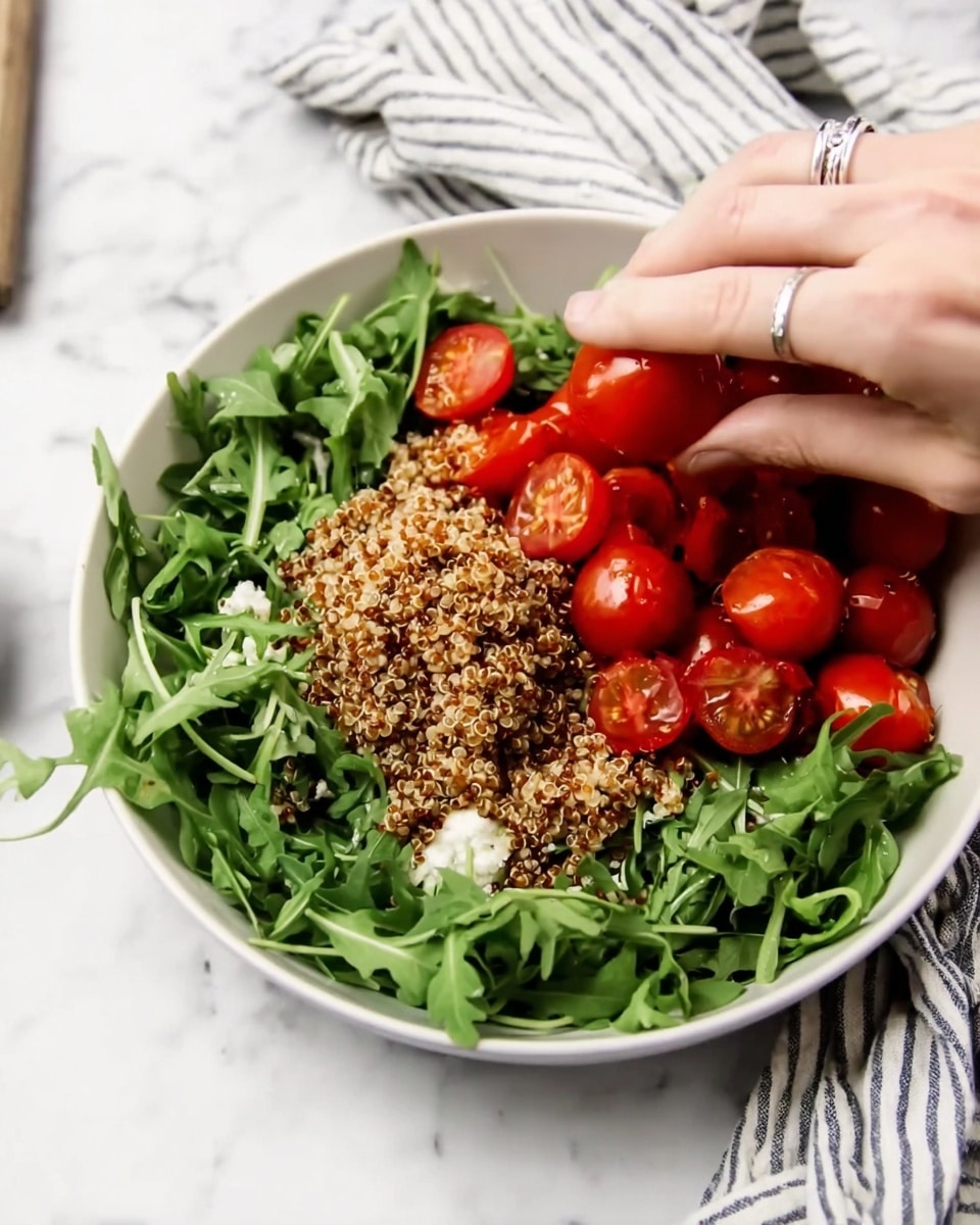 A white bowl sits on a white marbled surface filled with three main layers. The bottom layer is fresh green arugula leaves spread evenly in a loose bed. On top of this is a layer of cooked quinoa, textured with tiny grains and a light brown color, placed mostly in the middle. The top layer consists of bright red cherry tomatoes, some whole and some halved neatly on one side. A woman's hand with a silver ring is gently adjusting the tomatoes in the bowl. A striped cloth is blurred in the background. Photo taken with an iphone --ar 4:5 --v 7