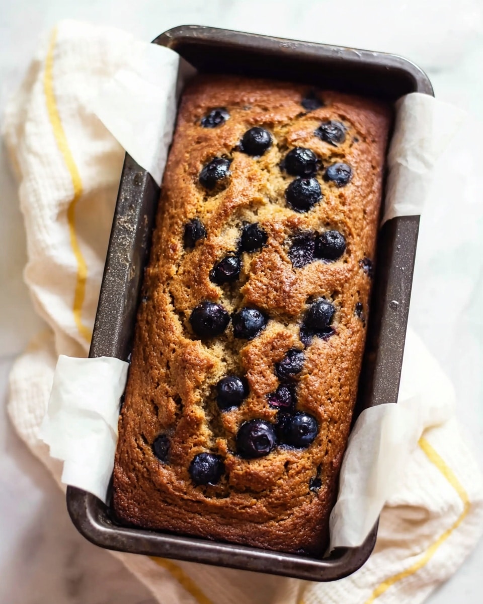 A golden brown baked loaf with a rough texture sits inside a dark metal rectangular pan lined with white parchment paper. Scattered across the top are plump, dark blue blueberries partially sunken into the bread, providing contrast to the warm brown crust. The surface shows some cracks and a slightly uneven rise, giving it a homemade look. The pan rests on a soft white cloth with light yellow stripes, all set on a white marbled surface. photo taken with an iphone --ar 4:5 --v 7