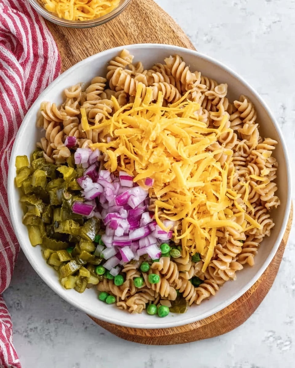 The dish is served in a white bowl filled with three main layers arranged distinctly. The bottom layer consists of light brown rotini pasta with small green peas mixed in, covering most of the base. On one side of the bowl, there are chopped green pickles with a slightly glossy texture. In the center, finely chopped red onions are spread over, creating a contrast with the other ingredients. On top of the onions, there is a bright yellow layer of shredded cheddar cheese that adds a pop of vibrant color. The bowl rests on a small wooden board placed on a white marbled surface with a red and white striped cloth nearby. Photo taken with an iphone --ar 4:5 --v 7