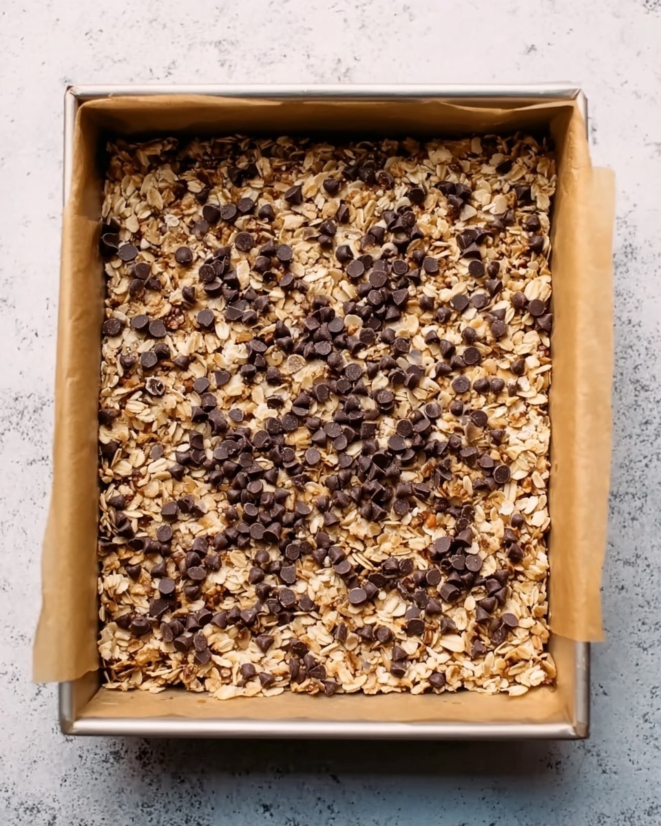 A square baking pan lined with brown parchment paper holds a single layer of a granola mixture. The granola layer is light golden brown with visible oats and small chunks of nuts creating a rough, textured surface. Dark chocolate chips are sprinkled unevenly but generously across the top, contrasting sharply with the lighter oats beneath. The pan sits on a white marbled textured surface. photo taken with an iphone --ar 4:5 --v 7