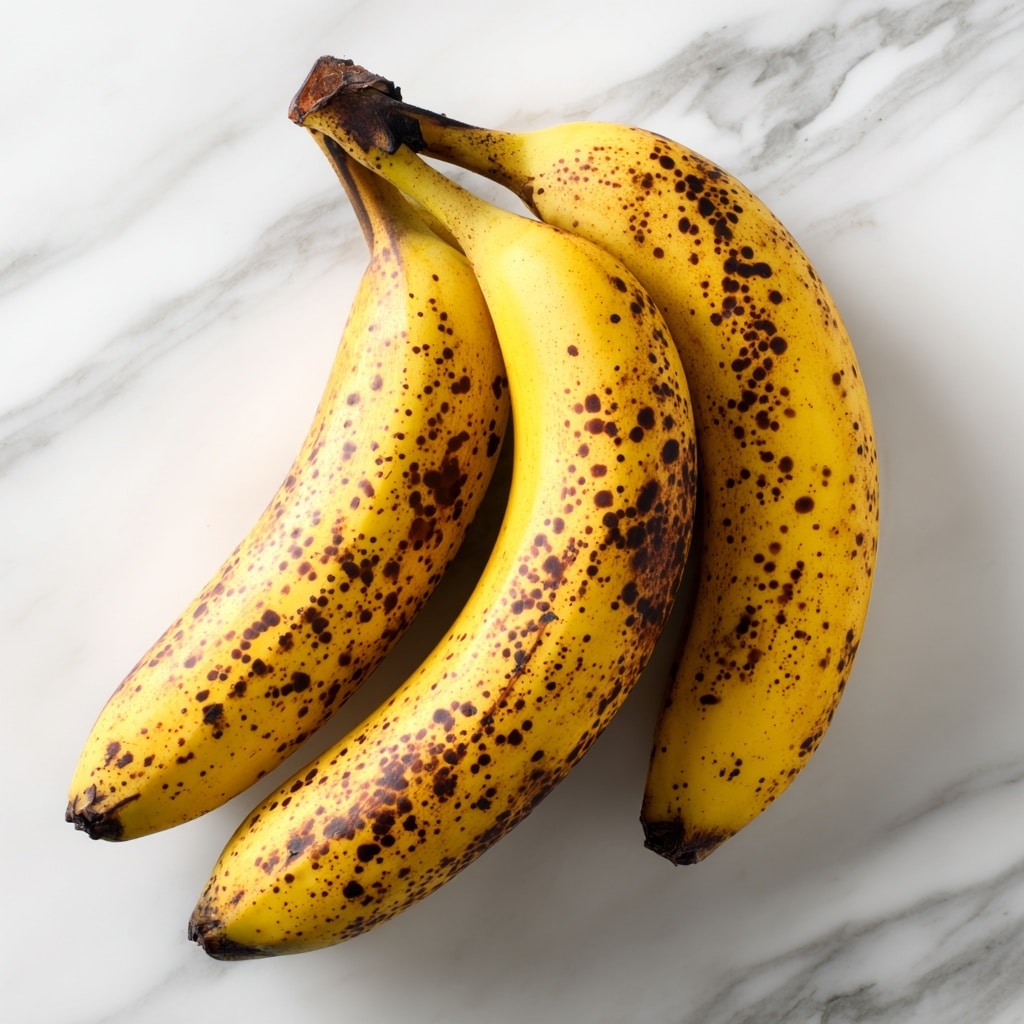 A cluster of three ripe yellow bananas with brown spots and streaks is shown from above, with the stems connected at the top left. The bananas have a slightly curved shape and a smooth texture with darkened ends, placed on a white marbled surface that adds a soft pattern in the background. The lighting is bright and natural, highlighting the bananas' color and spots photo taken with an iphone --ar 4:5 --v 7