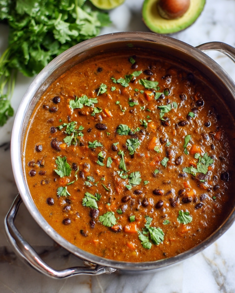 The image shows a pot filled with thick brown bean soup with small bits of black beans and orange specks throughout, suggesting the presence of finely chopped vegetables or spices. The soup’s texture looks creamy but still chunky, and it is garnished with small, bright green cilantro leaves scattered on top. The pot is metal with a sturdy handle visible on the side, and it sits on a white marbled surface. In the background, some green leafy vegetables and a partially cut avocado can be seen out of focus. Photo taken with an iphone --ar 4:5 --v 7