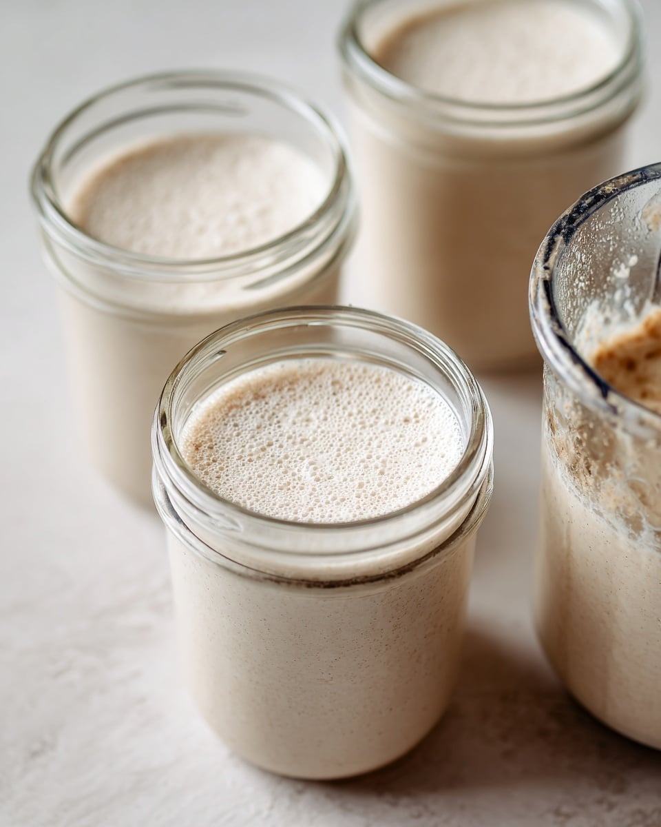 The image shows three clear glass jars filled with a light beige, creamy mixture, each almost full and slightly frothy on top. They are arranged closely together on a white marbled surface. To the right of the jars, there is a blender container with the same mixture inside, showing a smooth, slightly thick texture with bits visible near the top. The background is softly blurred to keep focus on the jars and blender. photo taken with an iphone --ar 4:5 --v 7