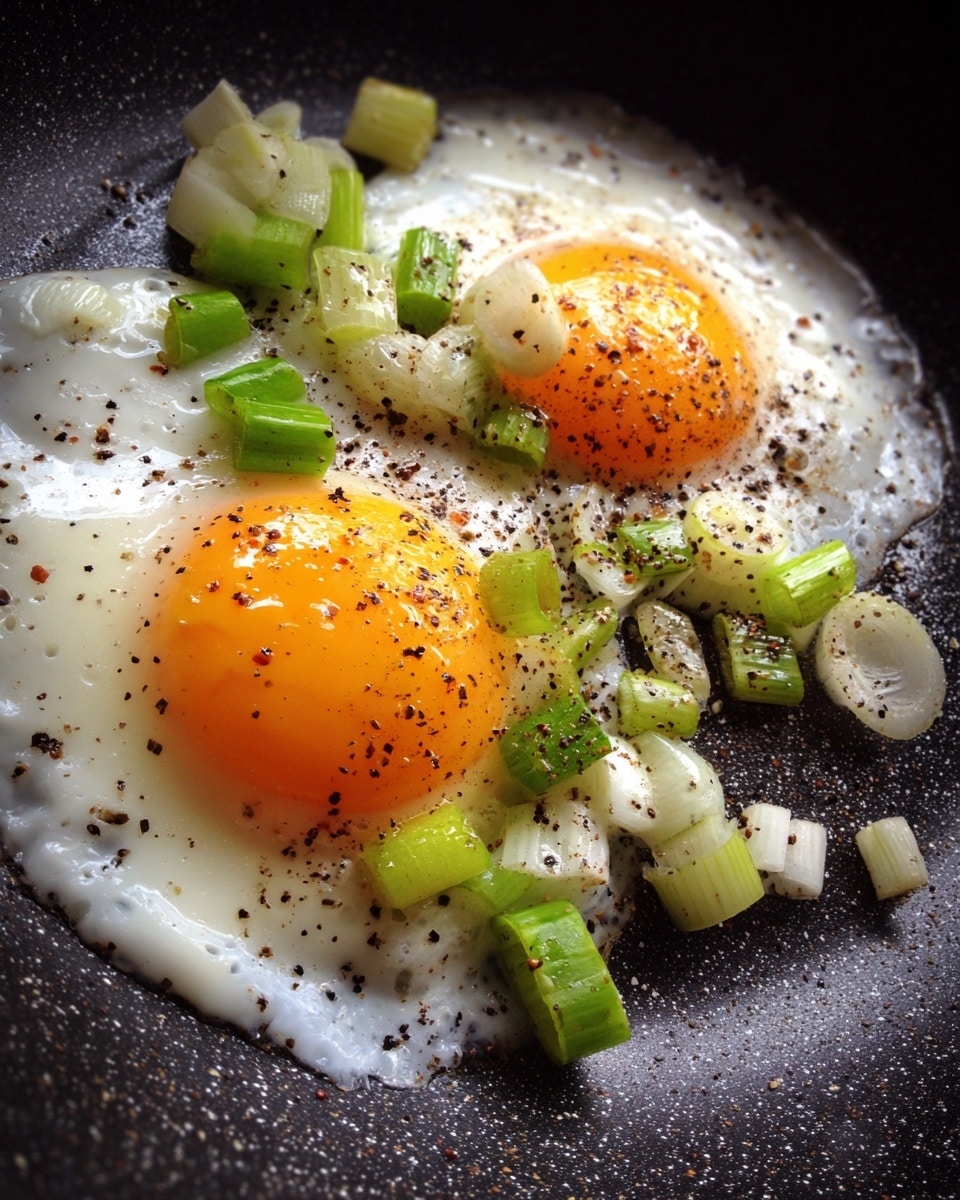 The image shows two bright orange cooked egg yolks surrounded by white cooked egg whites in a frying pan. Scattered on top and around the eggs are green chopped stems and slices of light green and white onion, with some black pepper sprinkled over. The background is a black speckled frying pan with a lightly shiny texture. photo taken with an iphone --ar 4:5 --v 7