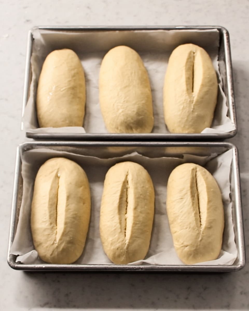 Six pieces of light beige dough with a smooth texture and a central score line are placed in two metal baking trays lined with white parchment paper. The trays rest on a white marbled surface, and the dough pieces are evenly spaced, three in each tray, ready for baking. The scene is simple and clean with warm lighting. Photo taken with an iphone --ar 4:5 --v 7