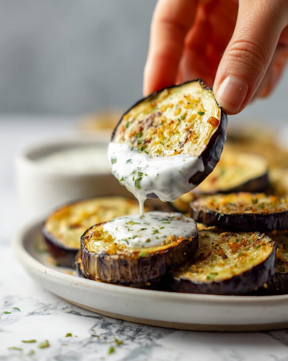 A close-up of a grilled eggplant slice held by a woman's hand, dipped in a thick white sauce with visible green herbs. The eggplant slice has a glossy, dark brown edge with a soft, golden-yellow center topped with small bits of seasoning, creating a textured surface. Below, there is a white plate holding more golden grilled eggplant slices with the same seasoning, placed on a white marbled surface. Photo taken with an iphone --ar 4:5 --v 7