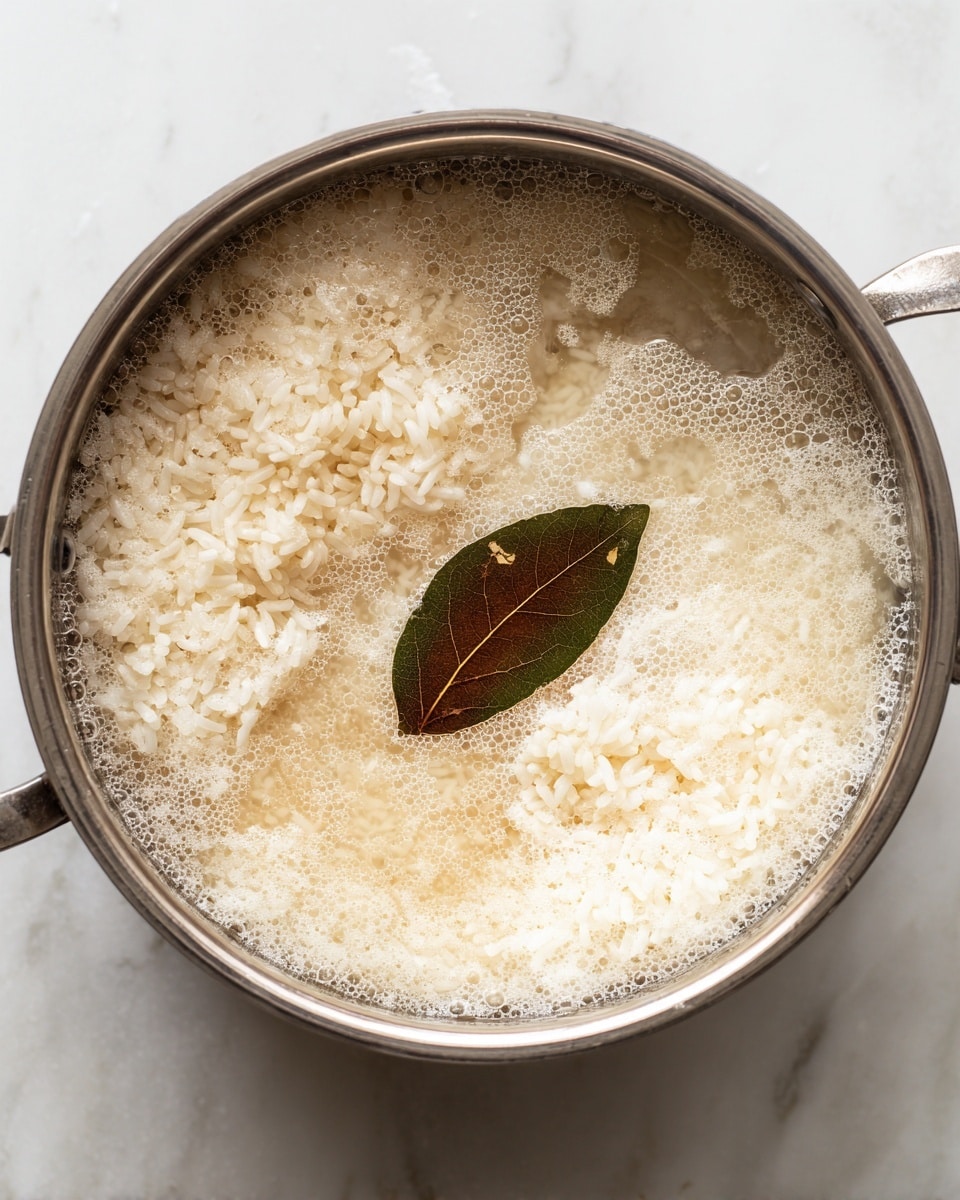 This image shows a pot with white rice cooking in clear water with bubbles on the surface, indicating it is boiling or simmering. In the middle of the rice, there is a single brown bay leaf floating on top. The water is slightly cloudy near the rice, and the texture highlights soft, individual rice grains partially submerged. The scene is viewed from above, focusing on the simple preparation stage of the dish, set against a white marbled texture background. photo taken with an iphone --ar 4:5 --v 7