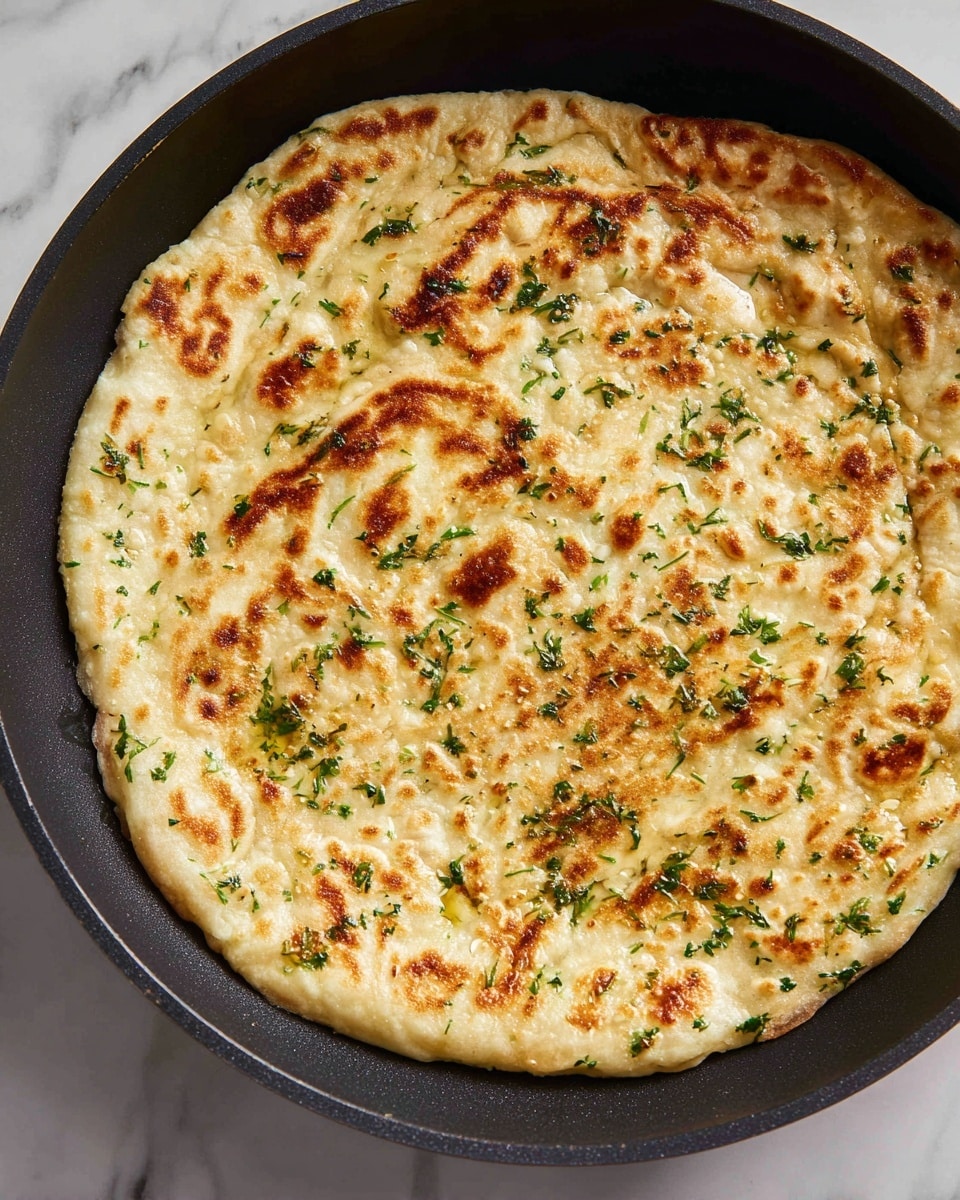 A single flatbread with small green herb bits spread throughout the dough, showing light golden brown spots from cooking, lies flat inside a black pan. The flatbread has a slightly soft and uneven textured surface with specks of green and golden brown scattered evenly, giving it a fresh and warm appearance. The black pan contrasts with the light color of the flatbread, making it stand out, with visible tiny oil droplets around the edge. The background is a white marbled texture. photo taken with an iphone --ar 4:5 --v 7