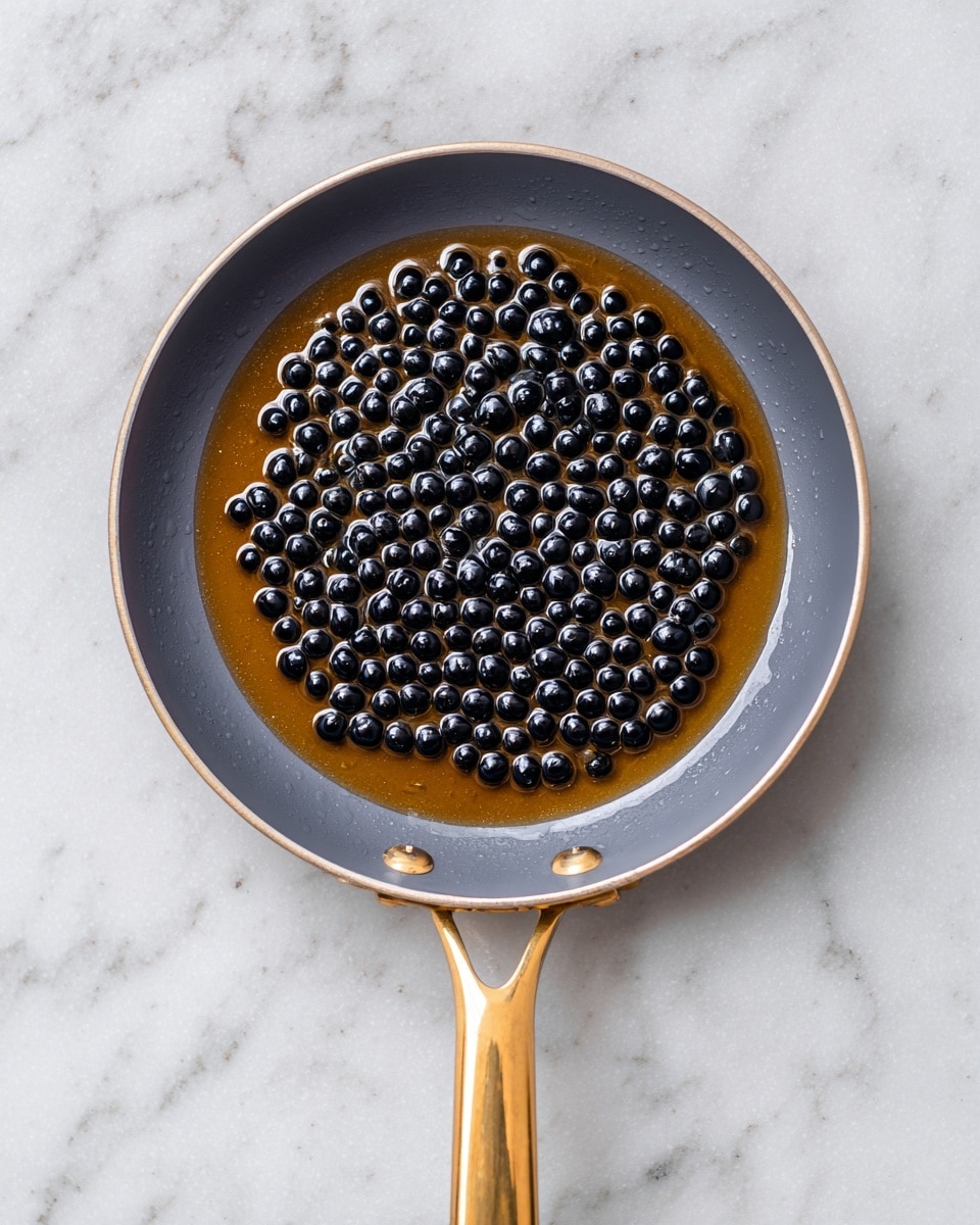 A gray frying pan with a golden handle holds a single layer of small, round, shiny black tapioca pearls soaked in a light brown syrup. The pearls cover the bottom of the pan evenly, with the syrup creating a glossy surface that contrasts with the matte gray interior of the pan. The background features a white marbled texture, enhancing the simplicity of the scene. photo taken with an iphone --ar 4:5 --v 7