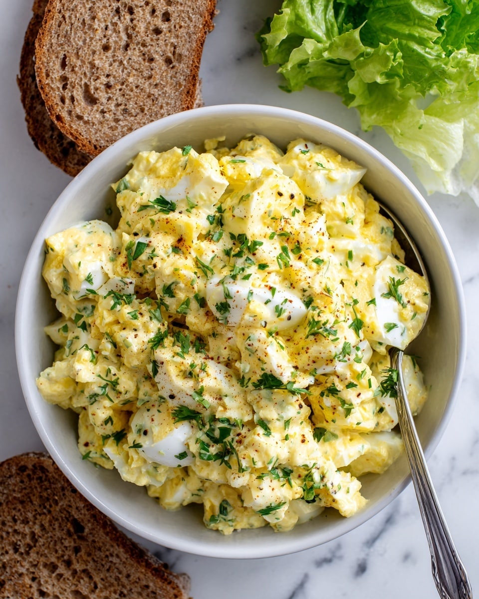 A close-up view of a white bowl filled with creamy egg salad, showing chopped hard-boiled eggs mixed with a yellow mayonnaise dressing, green herbs sprinkled on top, and visible bits of seasoning throughout. The bowl is placed on a white marbled surface, surrounded by three slices of whole grain bread on the left and fresh leafy lettuce at the top right. A metal spoon is partially submerged in the egg salad inside the bowl. Photo taken with an iphone --ar 4:5 --v 7