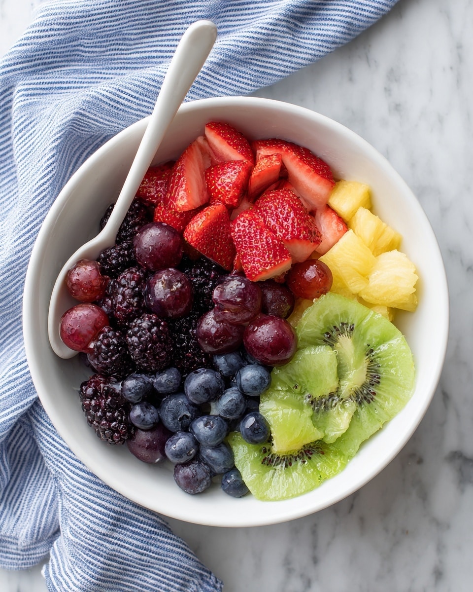 A close-up of a colorful fruit salad in a white bowl with many small fruit pieces visible. The layers show bright yellow pineapple chunks, green kiwi slices with black seeds, red strawberry halves with textured seeds, dark purple grapes, blueberries, and blackberries mixing together. A shiny silver spoon filled with a light, greenish dressing with small herb bits is being held above the fruit by a woman's hand. The background is a white marbled surface photo taken with an iphone --ar 4:5 --v 7