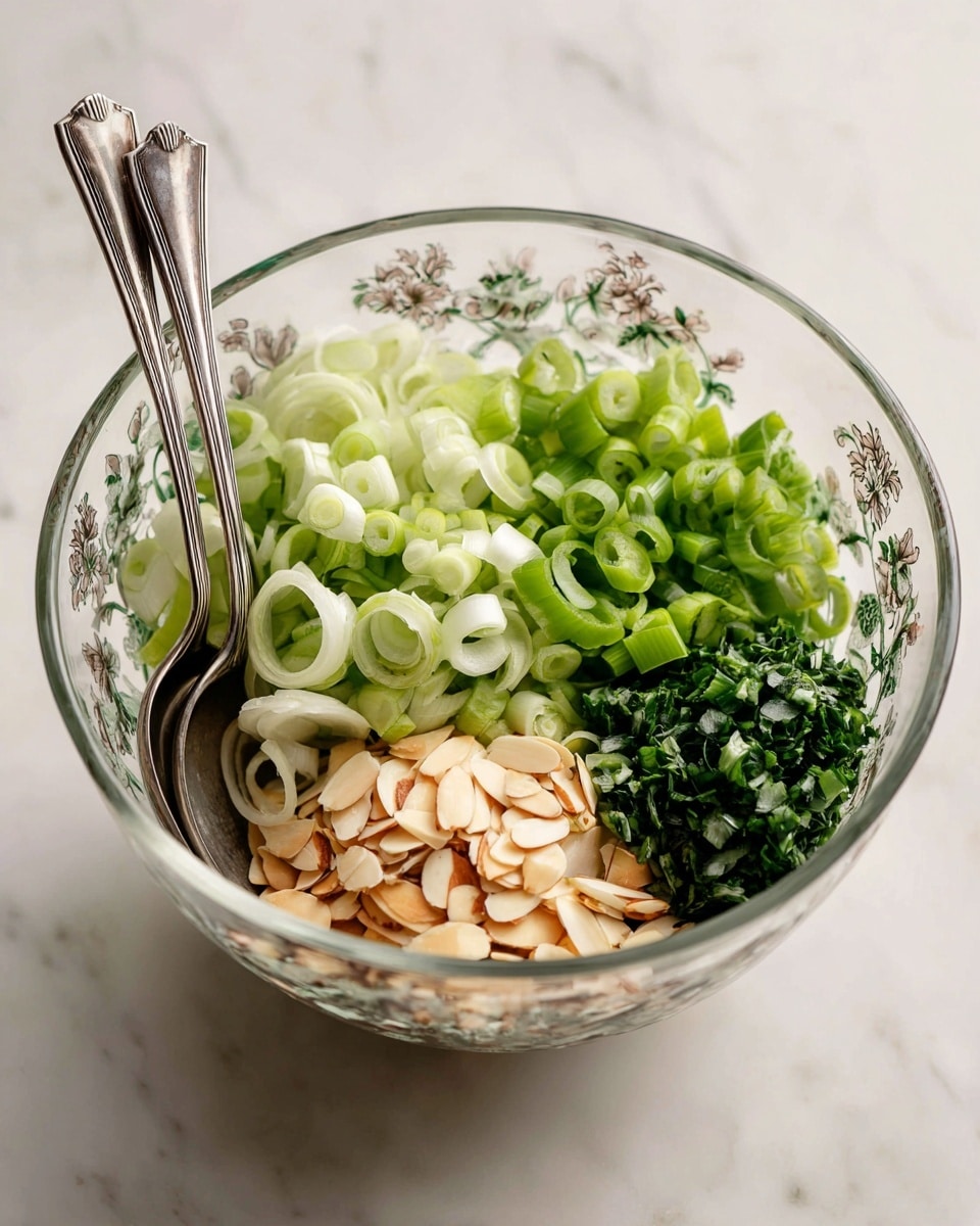 A clear glass bowl with floral designs holds fresh ingredients arranged in four sections on a white marbled surface. The top part has light green sliced celery, slightly curved and crisp in texture. Next to this are thinly sliced green onions with their pale green and white colors. To the right, there is a small pile of finely chopped dark green herbs, creating a dense leafy texture. At the bottom is a cluster of light brown almond slivers, smooth and slightly shiny. Two silver metal spoons rest inside the bowl on the left side, their handles stretching out. Photo taken with an iphone --ar 4:5 --v 7