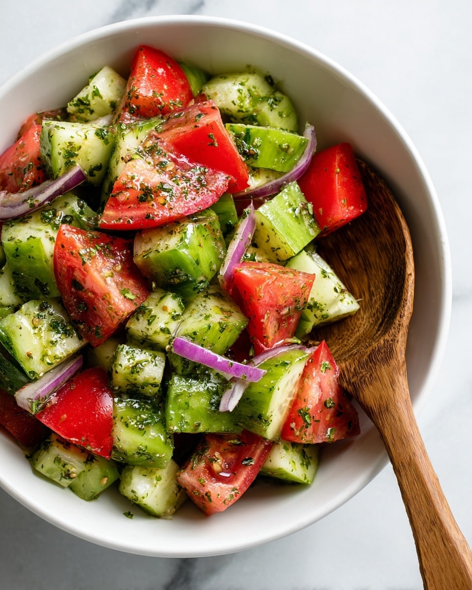 The image shows a fresh salad in a white bowl on a white marbled surface. The salad has three main layers: bright green cucumber pieces with a glossy texture scattered throughout, medium red tomato chunks with juicy, slightly shiny skin, and thin strips of purple-red onion spread evenly. The vegetables look lightly coated with a green herb dressing, with small bits of herbs sprinkled on the top. A wooden spoon with a smooth grain rests on the right side of the bowl, partially touching the salad, adding a natural touch to the composition. photo taken with an iphone --ar 4:5 --v 7