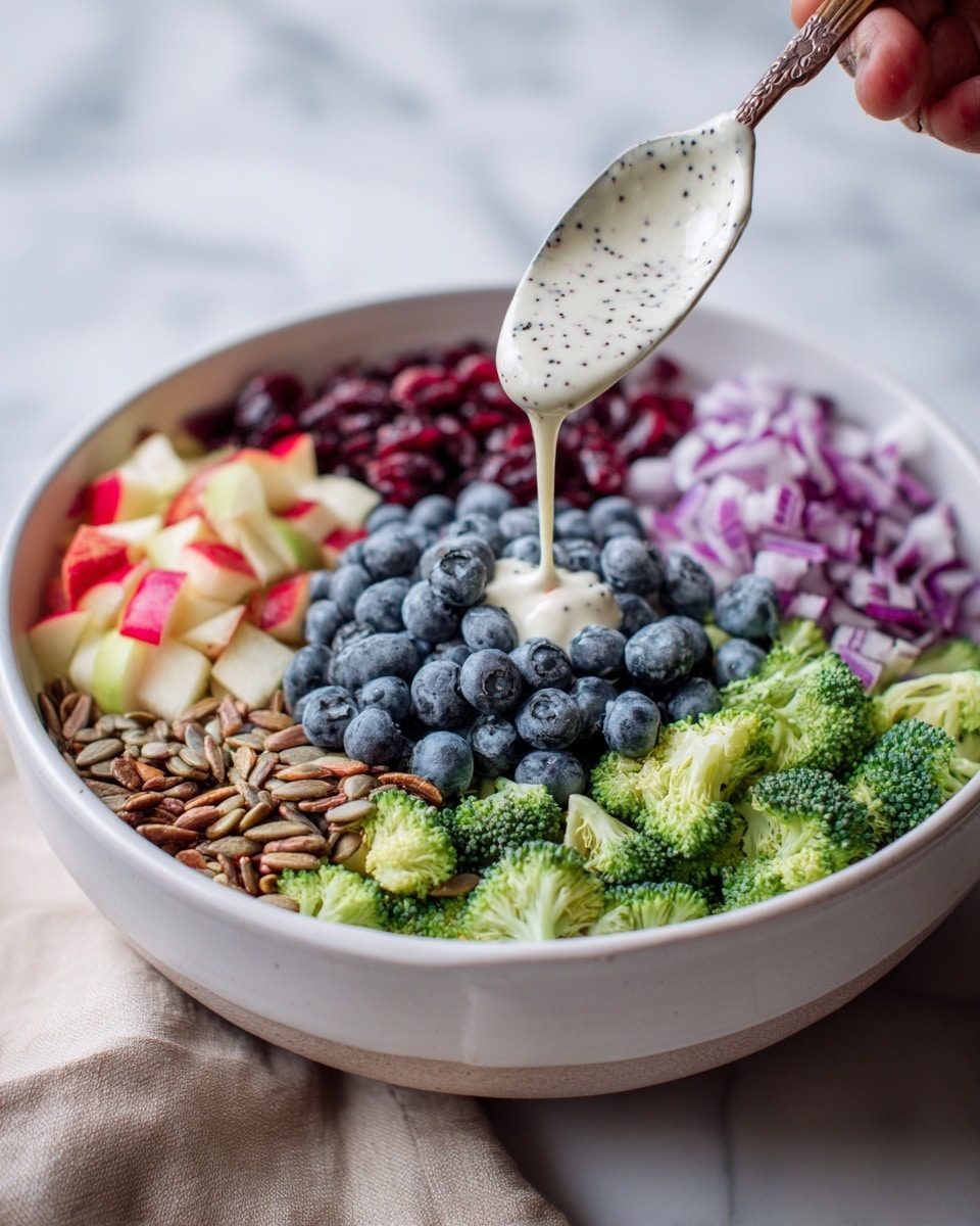 A white bowl sits on a white marbled surface filled with colorful layers of fresh salad ingredients. The bottom layer is green broccoli florets, topped with a bright layer of chopped red apples with white inside, next to a large pile of smooth, round blue blueberries. Beside the apples and blueberries, there is a sprinkling of light beige sunflower seeds and diced purple onions. Dried dark red cranberries add a pop of color next to the seeds. A woman's hand holds a white spoon dripping creamy, white dressing with small black seeds onto the center of the salad. Photo taken with an iphone --ar 4:5 --v 7