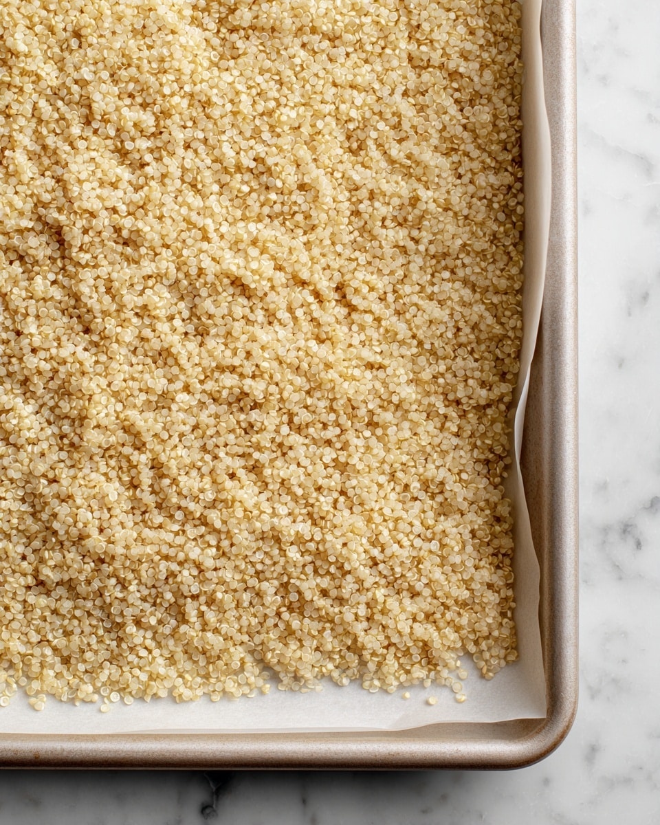 The image shows a single layer of light golden quinoa spread unevenly on white parchment paper, which itself is placed on a metal baking tray. The quinoa appears dry and toasted with some small clusters and slightly darker toasted edges scattered mostly around the perimeter. The background is a white marbled texture. photo taken with an iphone --ar 4:5 --v 7
