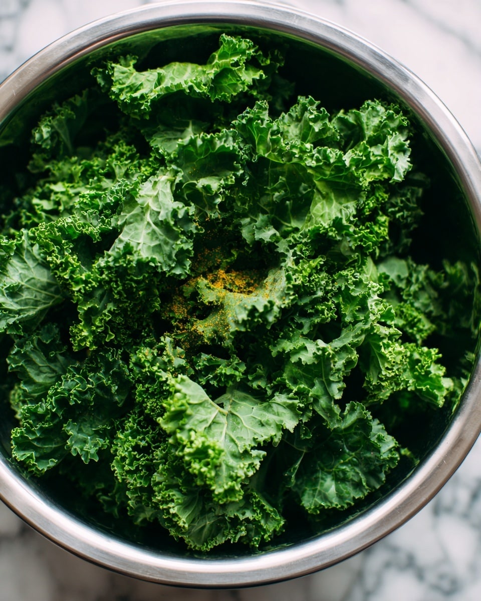 A close-up image of a large metal bowl filled with fresh, curly green kale leaves. The kale is bright green with detailed, ruffled edges, piled high and slightly overlapping in layers. In the center, there is a small sprinkle of orange powder on top of the kale, adding a small pop of color. The bowl sits on a white marbled surface with some background details blurred out. The photo has natural light highlighting the texture and freshness of the kale. photo taken with an iphone --ar 4:5 --v 7