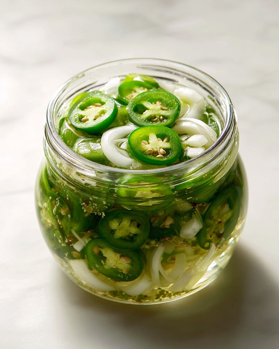 Inside a clear glass jar, there are two main layers visible from the top view. The bottom layer consists of thinly sliced green jalapeño peppers with their seeds showing, creating a bright and spicy green texture. Resting on top and mixed in between the jalapeños are thin, curved strips of white onions, adding a smooth and slightly translucent layer. The jar is filled with a clear pickling liquid that lets the green and white ingredients shine clearly. The jar is sitting on a white marbled surface. photo taken with an iphone --ar 4:5 --v 7