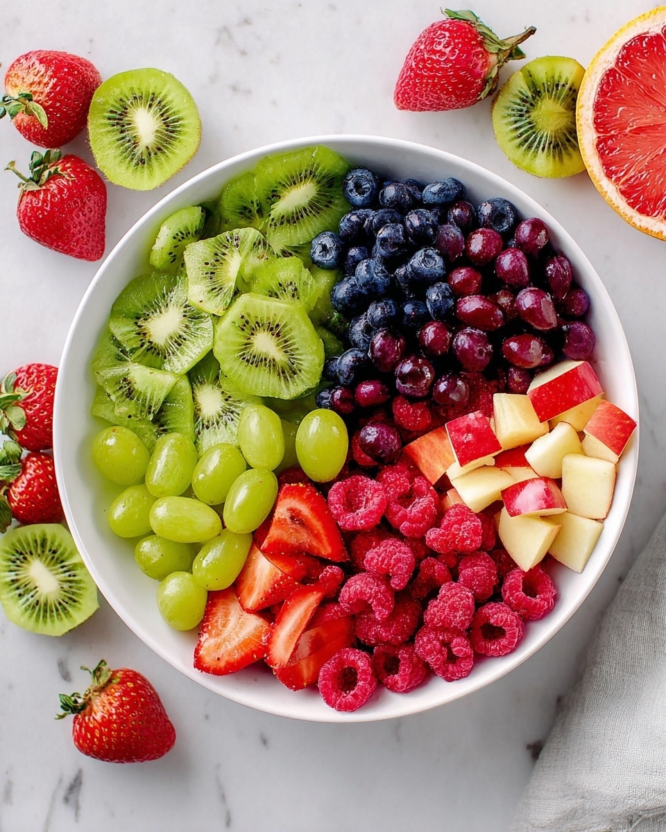 A white bowl filled with six types of fresh fruit arranged in separate sections: at the top left, thinly sliced bright green kiwi with a soft texture; to the right of the kiwi, dark purple blueberries, round and shiny; next to the blueberries, pieces of red apple with skin on, cut into small slices; below the apple slices, a pile of green grapes, some whole and some cut in half showing a juicy inside; to the left of the grapes, whole and sliced red strawberries with seeds visible; and at the far left bottom, a cluster of red raspberries with a bumpy texture. The bowl sits on a white marbled surface with scattered fresh fruits around it, including strawberries, kiwi halves, raspberries, grapes, and a halved grapefruit. Photo taken with an iphone --ar 4:5 --v 7