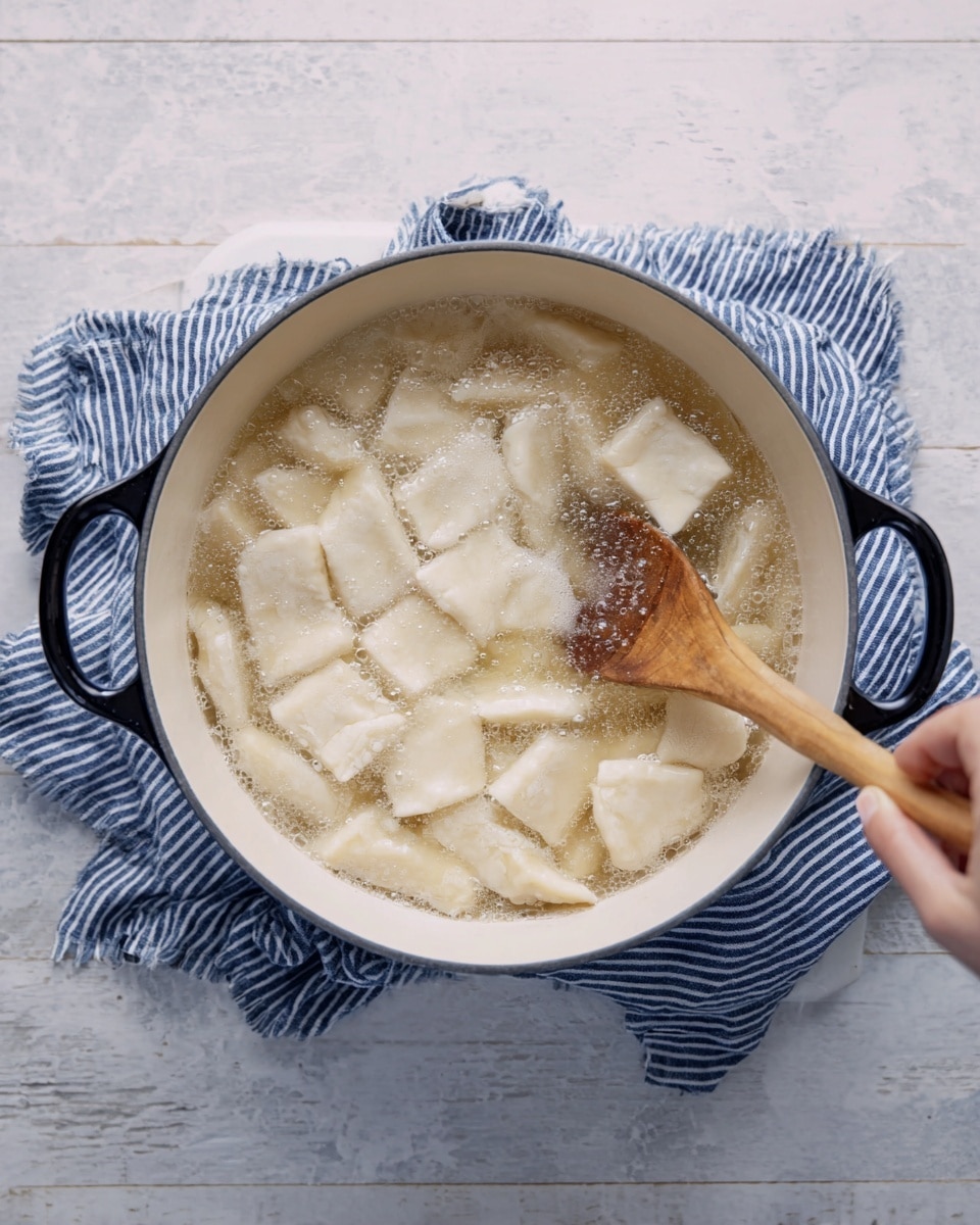 A white pot with black handles is filled with light beige dough pieces boiling in clear water, showing soft, uneven square shapes floating on the surface. A wooden spoon stirs the mixture from the right side, with a woman's hand holding the spoon near the bottom right corner. The pot rests on a blue and white striped cloth placed on a white marbled textured surface. photo taken with an iphone --ar 4:5 --v 7
