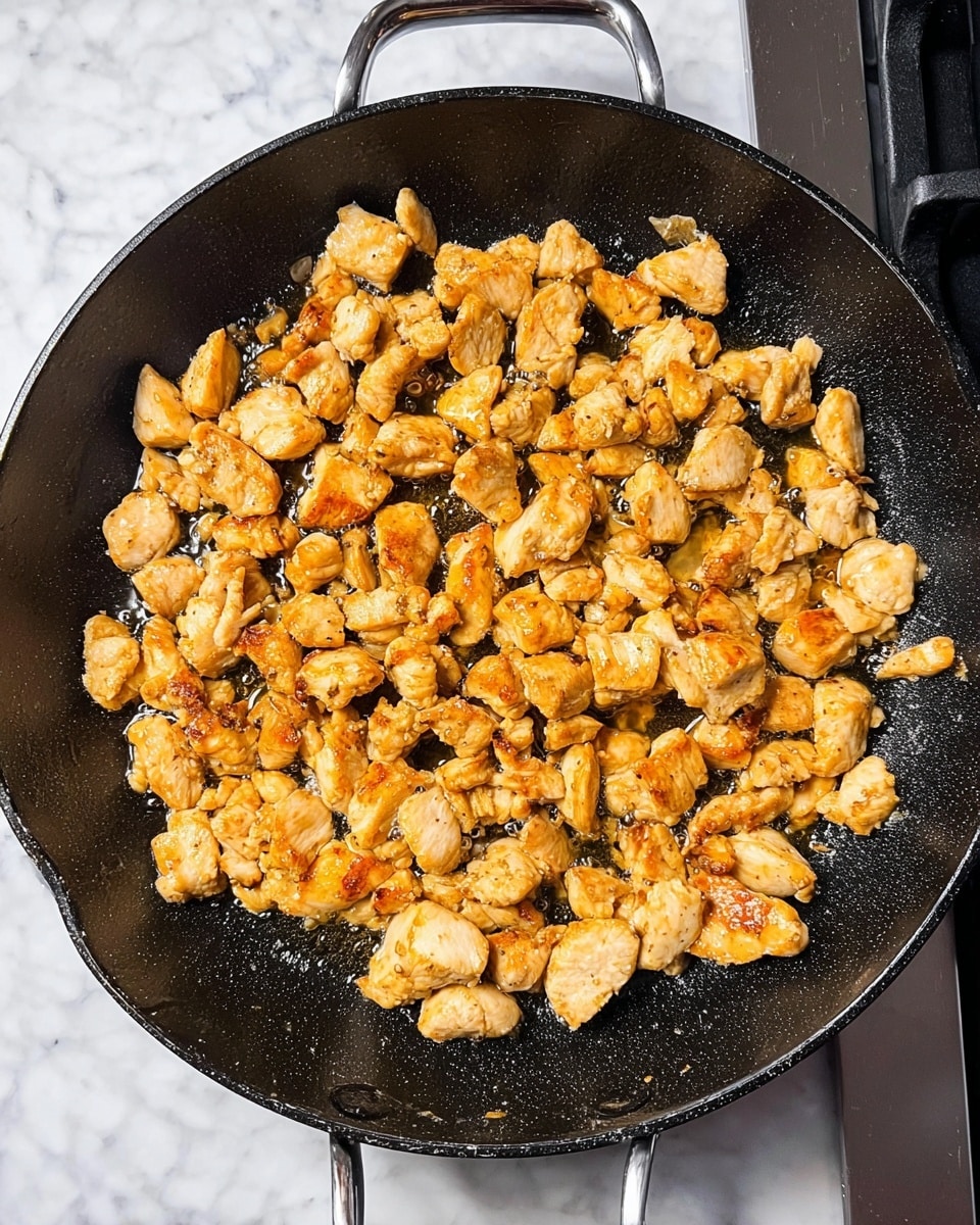 A close-up view of a black cast iron pan filled with many small pieces of cooked light golden brown chicken, scattered evenly across the pan's surface. The chicken pieces have a slightly crispy texture on the edges with a moist inside, and there is a small amount of sizzling oil surrounding them, giving a shiny look. The pan sits on a white marbled surface, with the handle of the pan visible at the top and bottom. The background shows a white marbled wall behind the pan. photo taken with an iphone --ar 4:5 --v 7