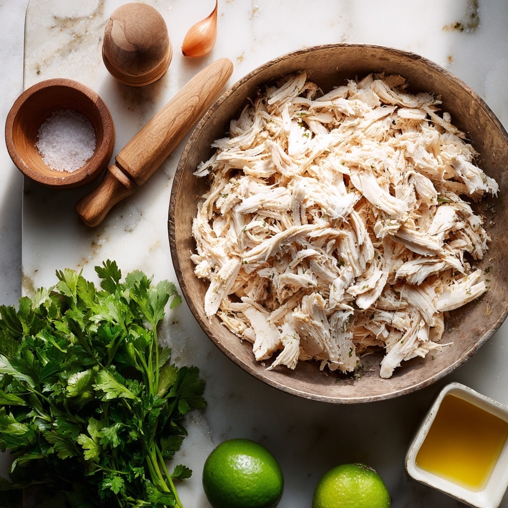 A large metal bowl filled with many shredded white cooked chicken pieces, showing soft texture with some darker bits; near it are two small shallow wooden salt containers, one open showing sea salt inside; two small light brown shallots sit next to a light wooden pestle, positioned diagonally; below, a bunch of fresh bright green cilantro or parsley with leafy textures, and a whole shiny green lime or lemon; at the bottom right, a small white square bowl holds golden olive oil, all items resting on a white marbled surface photo taken with an iphone --ar 4:5 --v 7