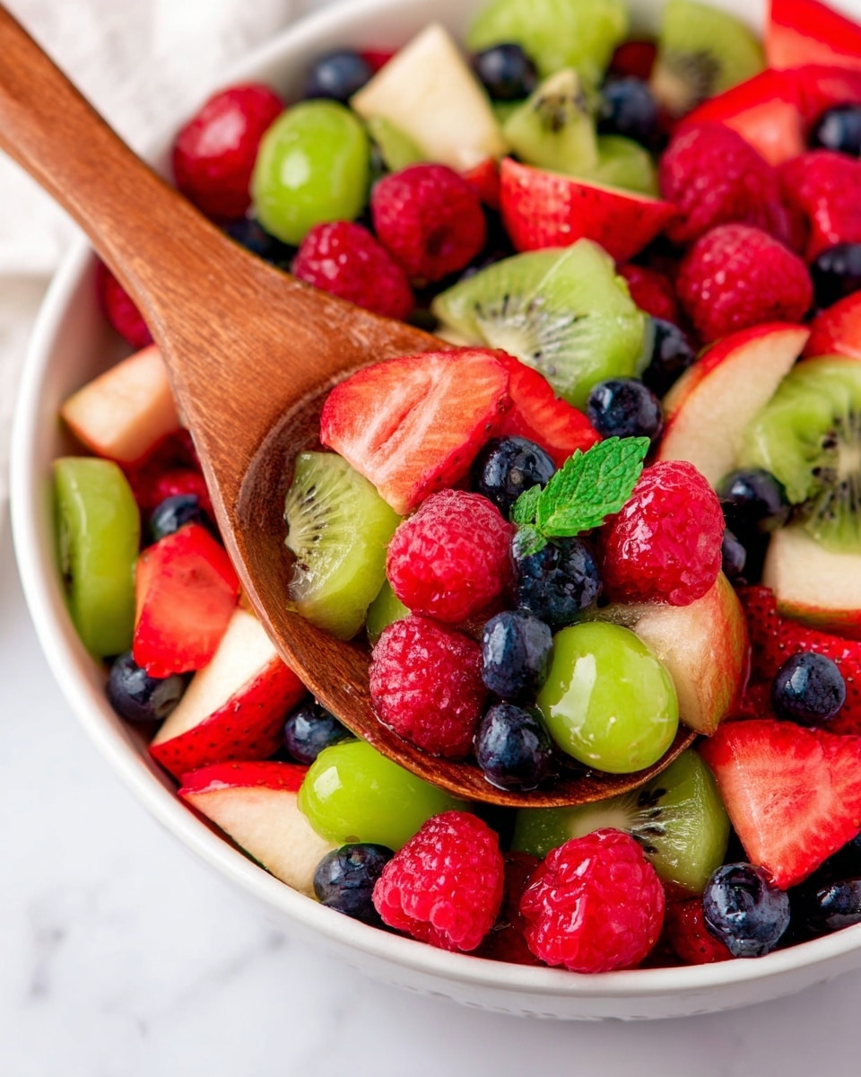A close-up of a fresh fruit salad in a white bowl on a white marbled surface, shown with a wooden spoon scooping some of the fruit. The salad has many colorful layers: bright red raspberries and strawberries, dark blue blueberries, light green kiwi slices, green grapes, and pale pink apple chunks with red skins. There is a small green leaf garnish on top. The wooden spoon’s texture is smooth and brown, resting inside the bowl near the center of the image. Photo taken with an iphone --ar 4:5 --v 7
