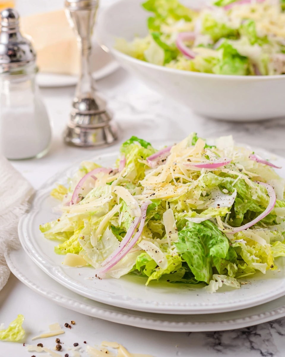 A white plate holds a fresh salad with several layers: the bottom layer is made of chopped green lettuce with a mix of light and dark shades, on top thin rings of pale purple onion are scattered. Small shreds of light yellow cheese are spread evenly over the salad, mixed with a few tiny pieces of black pepper. In the background, there is a larger white bowl filled with the same salad, placed on a white marbled surface with a salt shaker beside it. Some cheese shreds and bits of pepper are scattered around the plate. Photo taken with an iphone --ar 4:5 --v 7