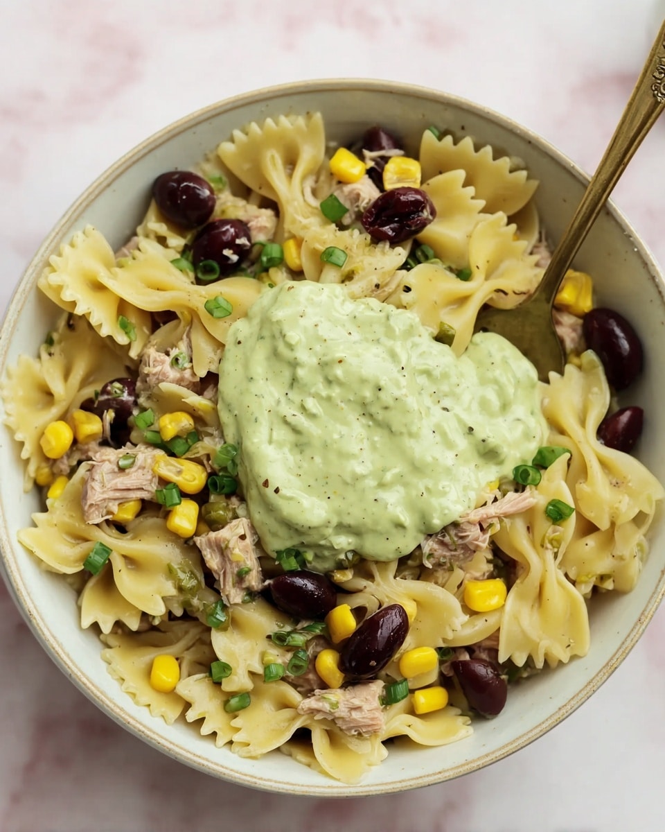 A white bowl filled with layers of small bow-tie pasta that are light yellow and smooth, mixed with chunks of light brown tuna, bright yellow corn kernels, dark purple olives, and small pieces of green onion scattered throughout. On top, there is a dollop of creamy green sauce with a slightly thick texture, and a spoon is partially dipped into the sauce and pasta inside the bowl. The bowl sits on a white marbled surface. photo taken with an iphone --ar 4:5 --v 7