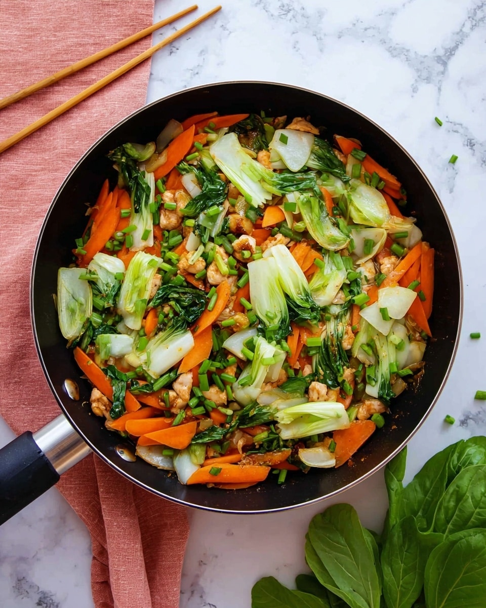 The image shows a black pan filled with a stir-fry mix of colorful ingredients. The bottom layer has bright orange carrot strips, cut roughly into long pieces. On top of this are light green and white bok choy pieces with a fresh, shiny texture. Scattered among the vegetables are small, lightly browned pieces of cooked chicken, adding a slightly crispy look. Small bits of green chopped chives are sprinkled evenly over the dish, enhancing the fresh feel. The pan is placed on a white marbled surface, and a pale pink cloth with wooden chopsticks lies near the pan's left side. A bunch of fresh green leaves is visible on the right side of the image. The photo taken with an iphone --ar 4:5 --v 7