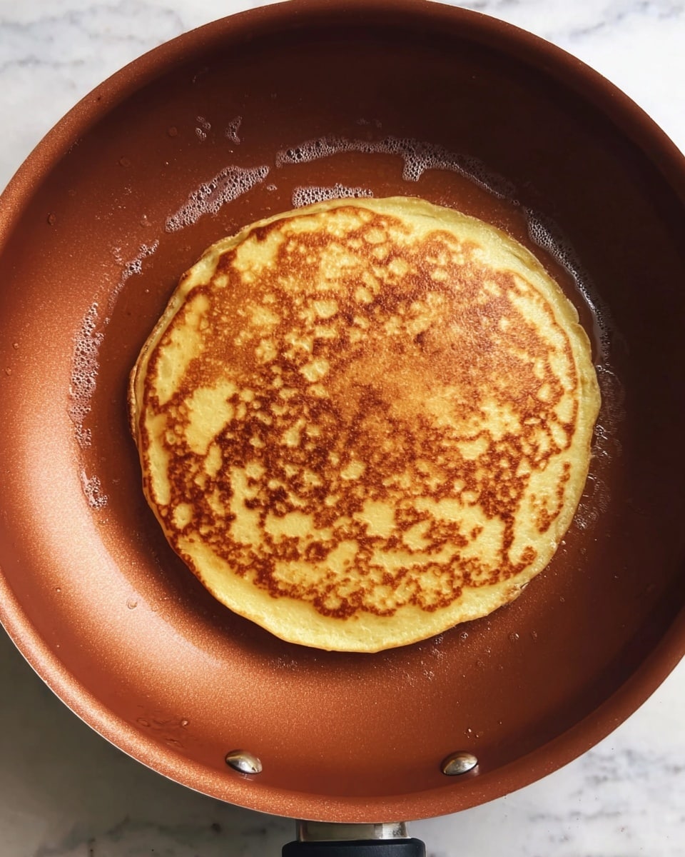 A single round pancake with a golden-brown surface and some darker brown spots is cooking in the center of a copper-colored non-stick frying pan. The pancake has a slightly uneven edge and looks fluffy with a smooth texture. Small bubbles of oil can be seen around the pancake inside the pan. The pan rests on a white marbled surface. photo taken with an iphone --ar 4:5 --v 7