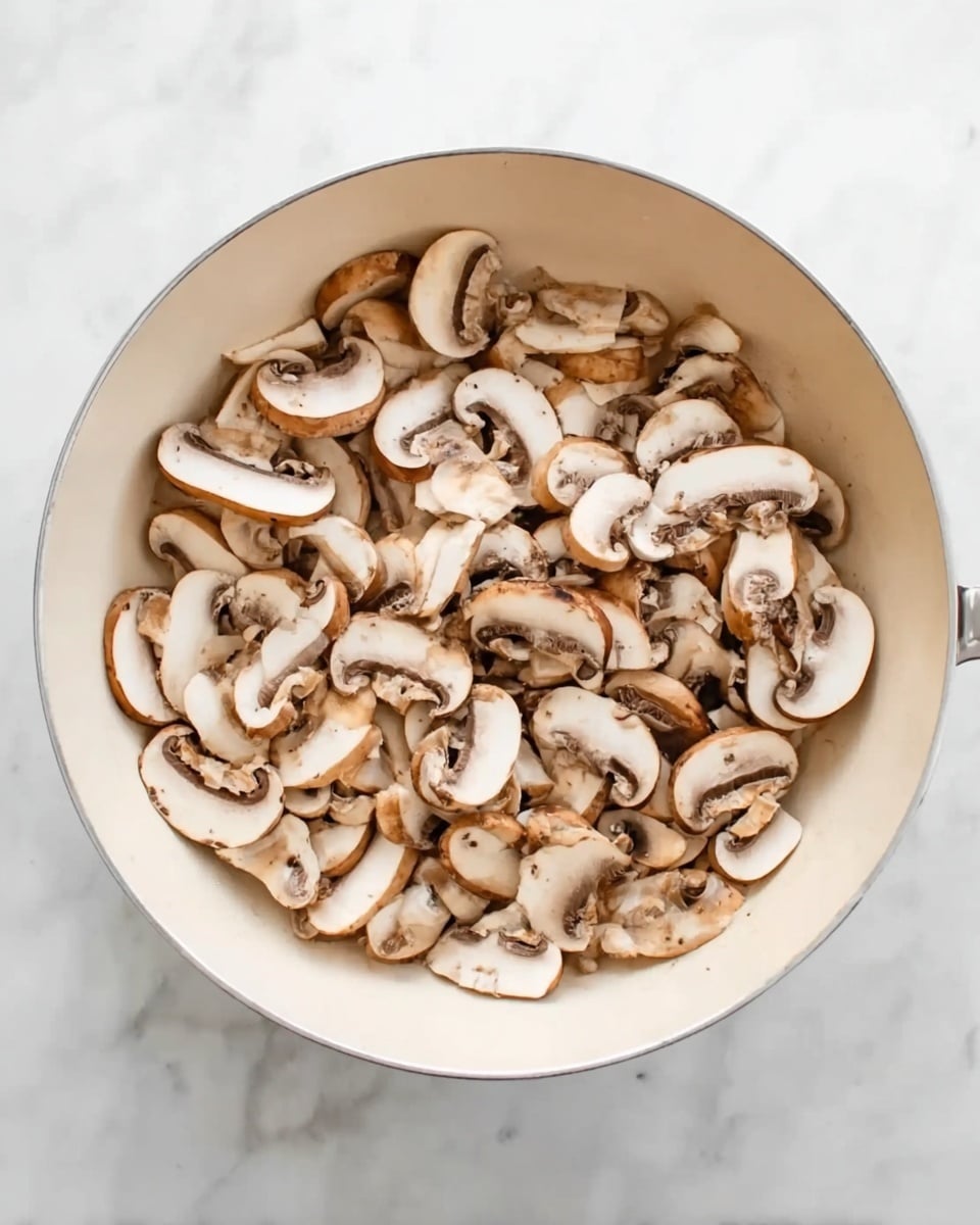 The image shows a white pan filled with many sliced mushrooms. The mushrooms are light brown and white, layered closely together, covering almost the whole surface inside the pan. The pan sits on a white marbled surface that serves as the background. The mushrooms have a soft texture with smooth edges and small dark spots in the middle where the stems are visible. There are no other ingredients visible in the pan. photo taken with an iphone --ar 4:5 --v 7