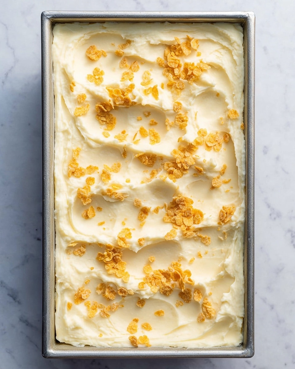 A rectangular metal pan filled with one smooth, creamy white layer of mashed potatoes spread evenly to the edges. The mashed potatoes have swirled textures with small peaks and valleys, topped with scattered golden-yellow crushed cornflakes for added crunch. The pan sits on a white marbled surface, giving a clean and bright background. photo taken with an iphone --ar 4:5 --v 7