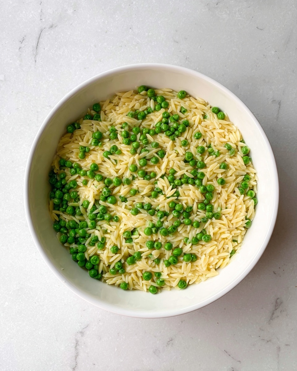 A white bowl filled with one layer of small, cooked orzo pasta in a light creamy yellow color mixed evenly with bright green peas. The orzo grains and peas are spread out in the bowl, creating a simple and fresh look. The bowl sits on a white marbled surface with soft natural light brightening the scene. photo taken with an iphone --ar 4:5 --v 7
