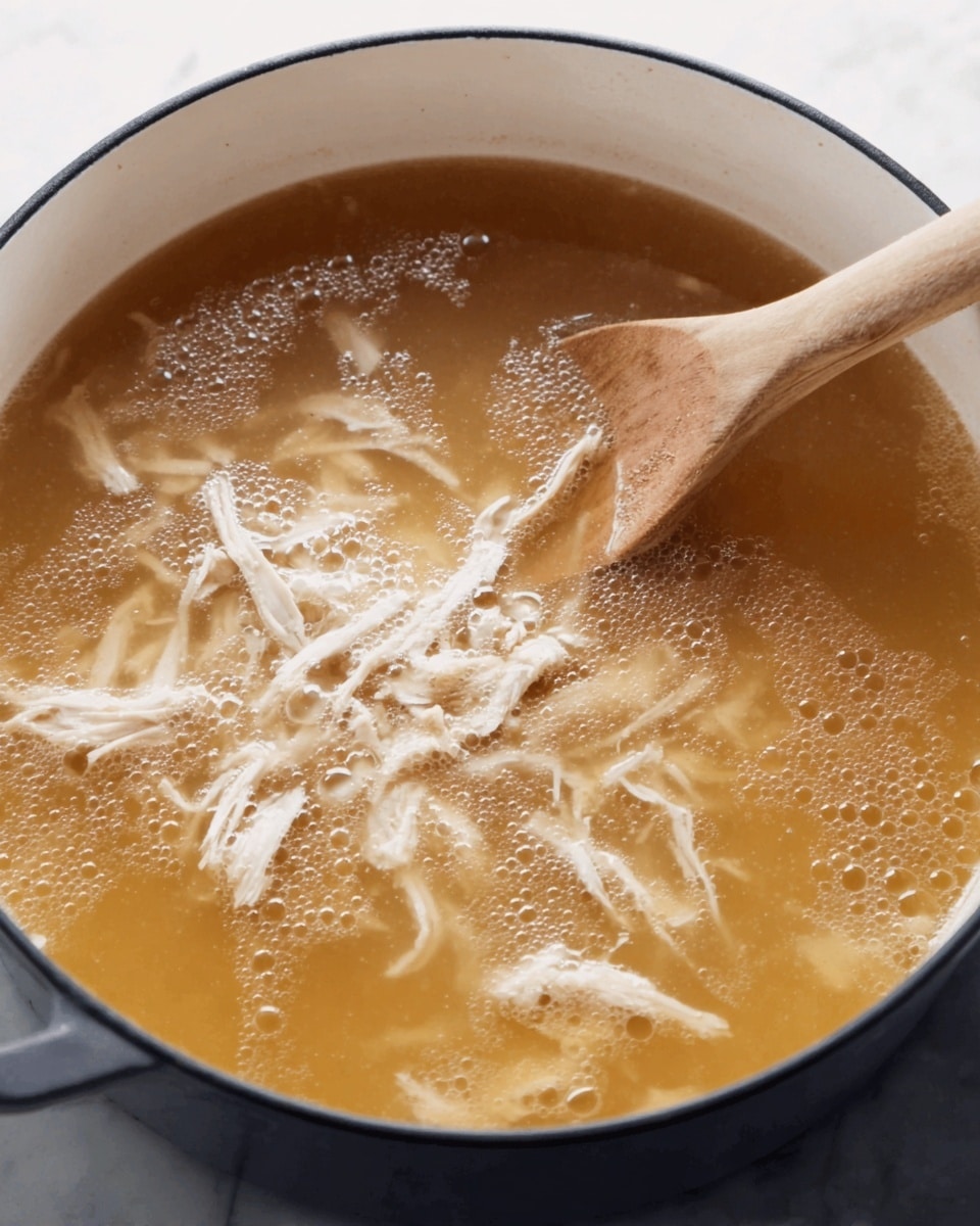 A close-up view of a pot with light brown broth and shredded white chicken meat floating on the surface. The pot is white inside with a dark rim, and a wooden spoon is stirring the broth from the right side. Small bubbles gather near the edges of the broth, giving a simmering effect. The setting is on a white marbled surface. photo taken with an iphone --ar 4:5 --v 7