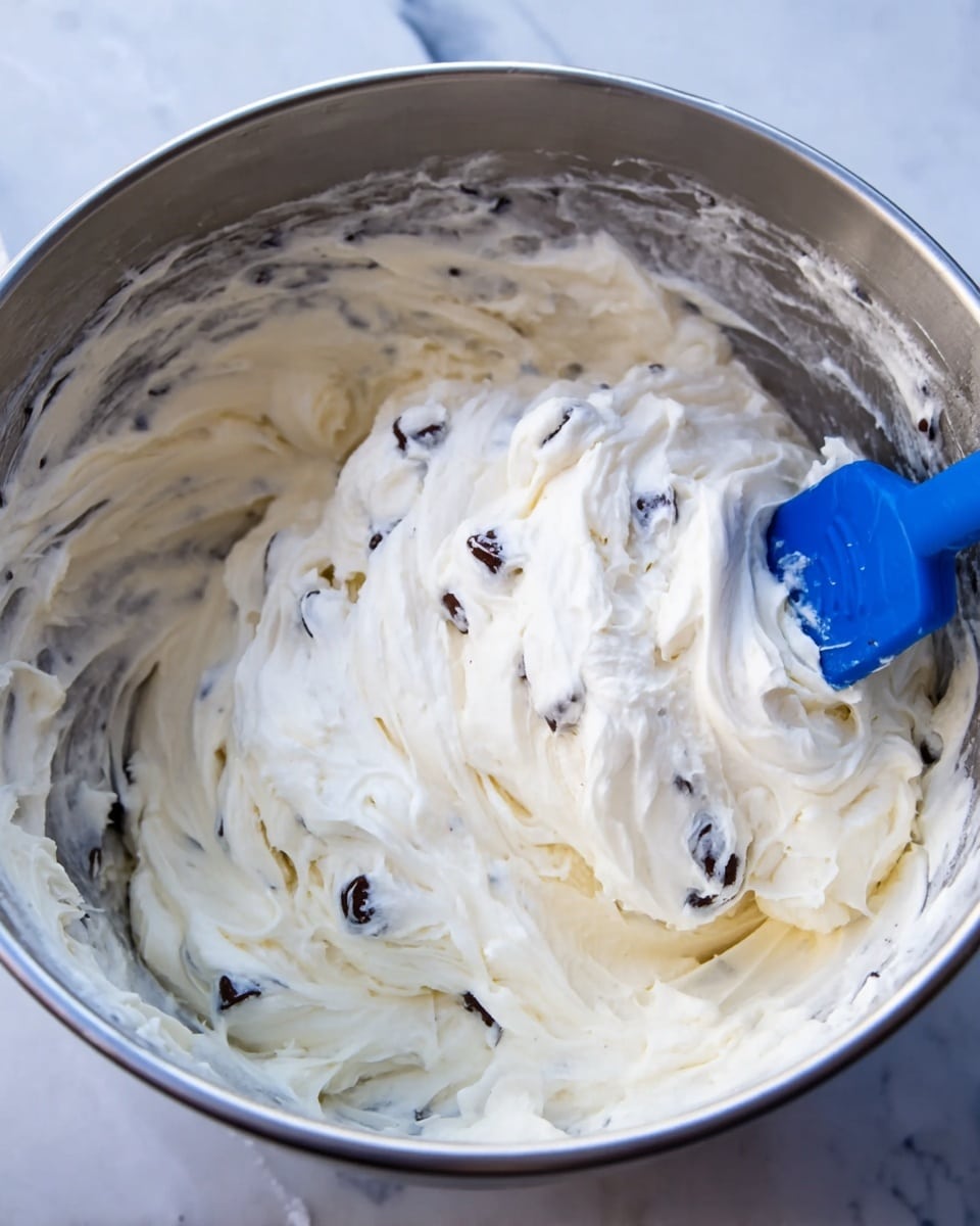 A close-up view inside a shiny silver mixing bowl shows a thick, creamy white batter with soft, smooth texture. Scattered evenly throughout are small, dark chocolate chips that stand out against the white batter. A blue spatula with a metal handle is partially submerged in the mixture, showing the creamy folds and swirls as it gently mixes the ingredients. The bowl sits on a white marbled surface, highlighting the creamy white batter and the contrast of the dark chips. photo taken with an iphone --ar 4:5 --v 7