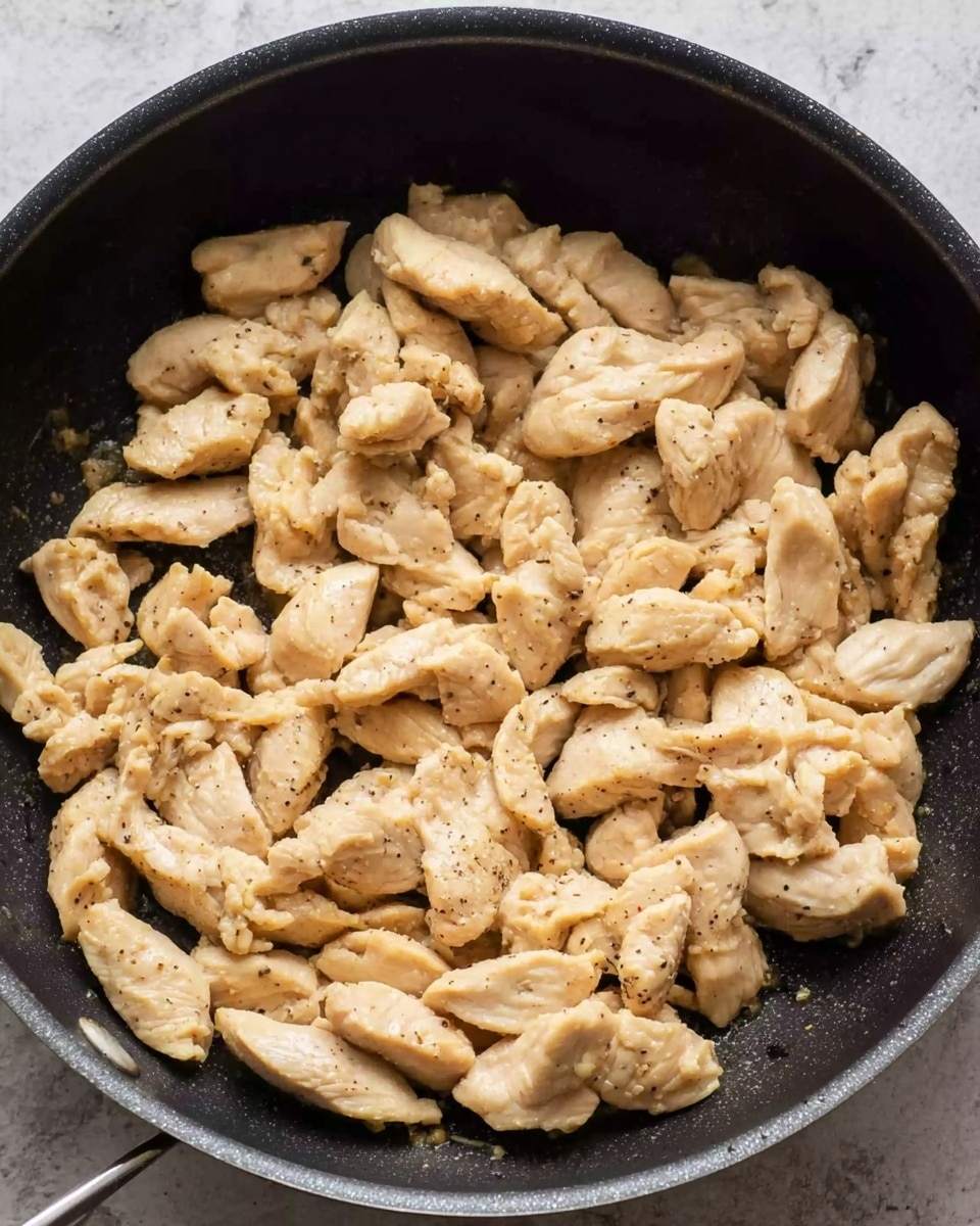The image shows a black pan filled with many pieces of cooked sliced chicken. The chicken pieces are light tan with some black pepper spots, unevenly scattered and layered inside the pan. The texture of the chicken looks tender and slightly moist, with some pieces curled or folded. The pan sits on a surface that is changed to a white marbled texture in the background. photo taken with an iphone --ar 4:5 --v 7