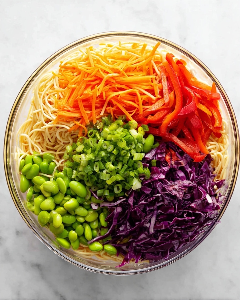 A clear round bowl on a white marbled surface holds a colorful layered noodle salad. The base layer is pale yellow thin noodles filling the bowl. On top, there are five vegetable layers arranged in sections: shredded bright orange carrots at the top left, thin red bell pepper strips at the top right, a pile of chopped green onions in the middle, vibrant green shelled edamame beans at the bottom left, and shredded deep purple cabbage at the bottom right. The colors are fresh and bright, creating a vivid mix. photo taken with an iphone --ar 4:5 --v 7
