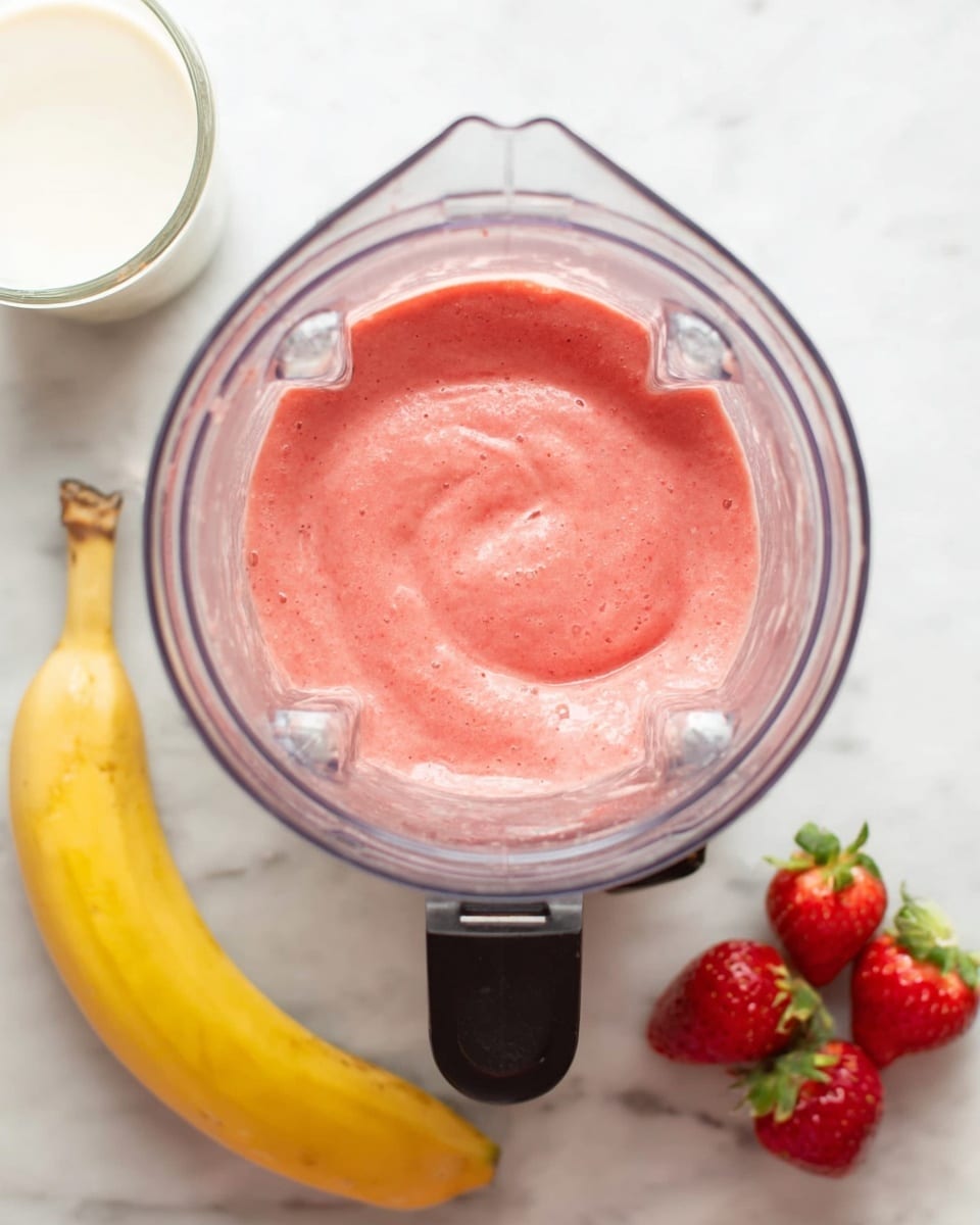 A top view of a blender filled with a thick, pink smoothie mixture that has a smooth and creamy texture. Just below the blender at the bottom of the image, there is a yellow banana on the left side and a cluster of three bright red strawberries with green leaves on the right side, all placed on a white marbled surface. To the left, there is a small white glass of milk or yogurt visible. The blender is clear plastic with a black base, centered on a white marbled background. Photo taken with an iphone --ar 4:5 --v 7