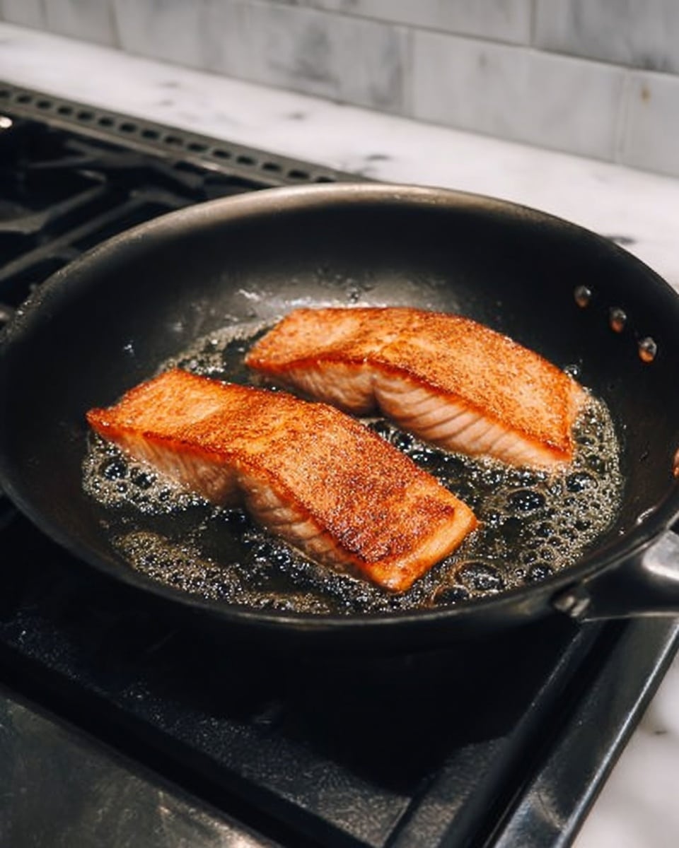 Two pieces of salmon are frying in a black pan on a stove. The salmon fillets have a light orange color with crispy brown edges from cooking. The pan has some oil that shimmers and bubbles around the fish. The stove has black metal grates and white tiled walls in the background. The surface below the stove is white marble. photo taken with an iphone --ar 4:5 --v 7