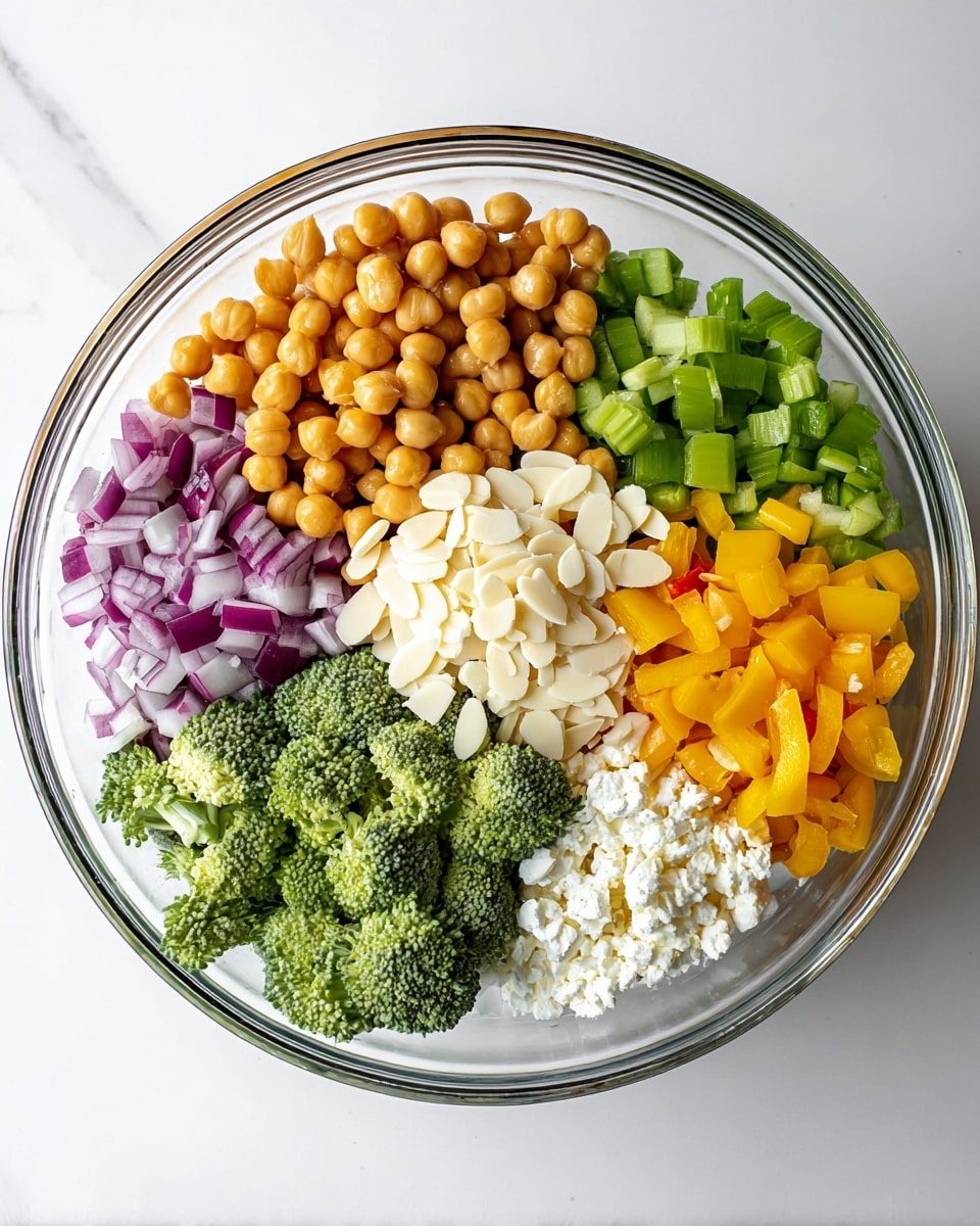 A clear glass bowl sits on a white marbled surface, holding six layers of ingredients arranged separately: golden chickpeas at the top left, purple-red chopped onions at the top right, light green chopped celery at the middle right, bright yellow diced bell peppers at the bottom right, white crumbly cheese heaped near the center, and fresh dark green broccoli florets at the bottom left. Thin white slivered almonds rest in the center on top of the cheese, adding texture and contrast. The bowl is viewed from above with bright, even lighting showing the fresh colors clearly. photo taken with an iphone --ar 4:5 --v 7