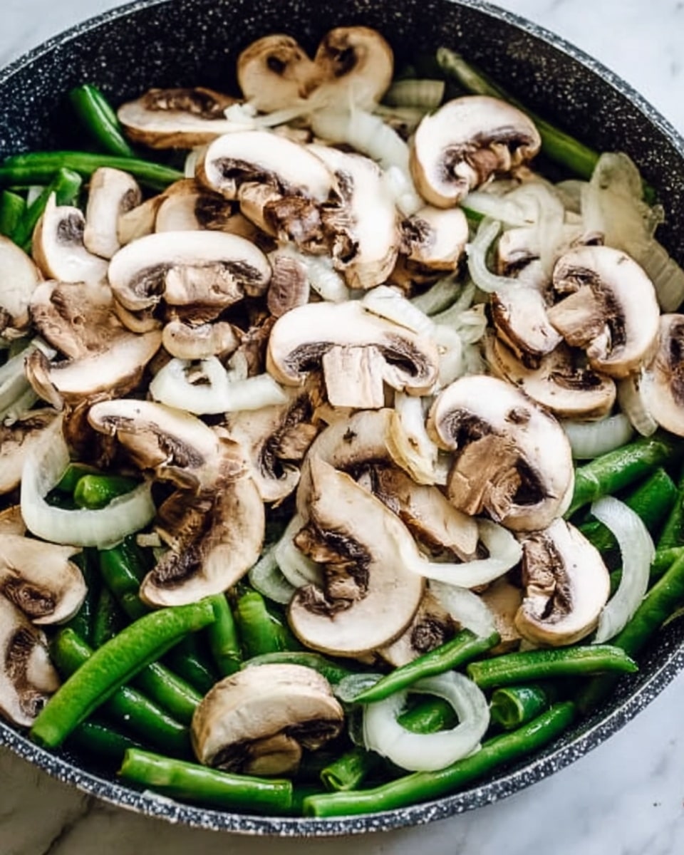 A close-up view of a pan filled with two layers: the bottom layer is green beans with white onion slices scattered among them, both fresh and vibrant; the top layer is sliced white mushrooms with light brown gills and stems, covering the green vegetables evenly; the pan has a dark rim and speckled texture, placed on a white marbled surface. photo taken with an iphone --ar 4:5 --v 7