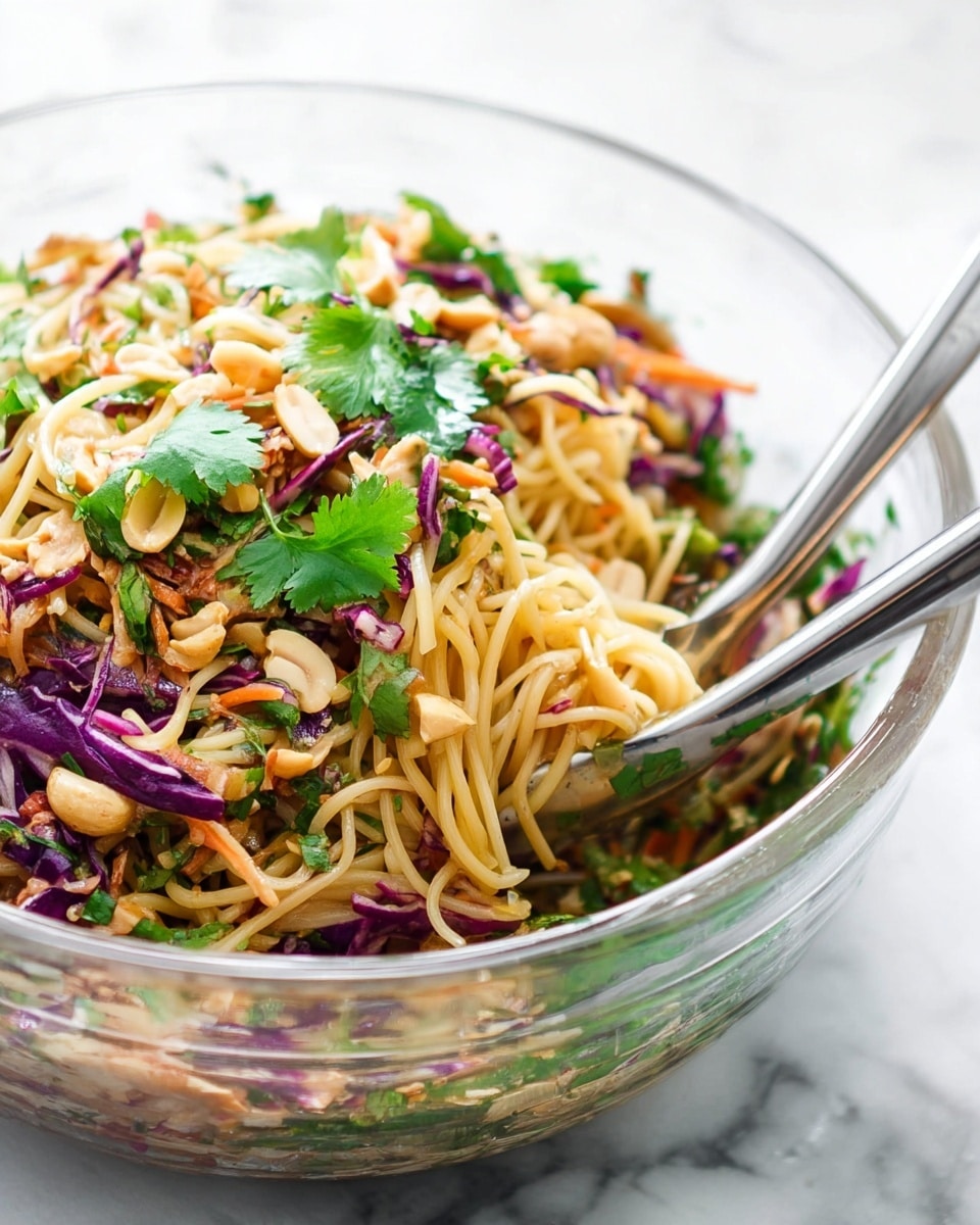 A clear glass bowl filled with a colorful noodle salad sits on a white marbled surface. The salad has three main layers: a bottom layer of mixed green herbs including cilantro, a middle layer of light yellow noodles, and a top layer scattered with shredded purple cabbage, light brown peanuts, small carrot pieces, and fresh green cilantro leaves. The textures are fresh and crunchy with a mix of soft noodles and crisp vegetables. Silver metal tongs are placed inside the bowl for serving. Photo taken with an iphone --ar 4:5 --v 7