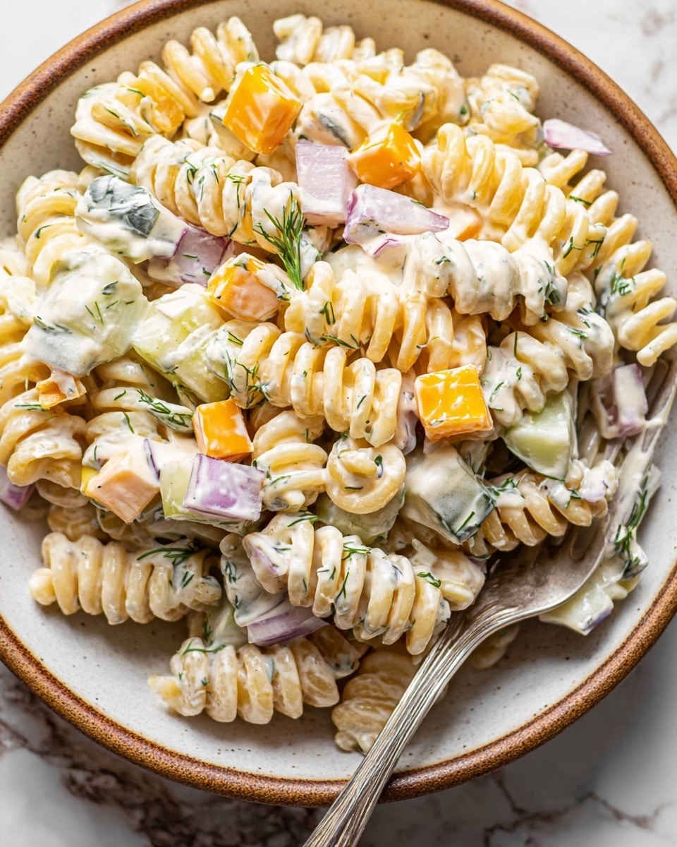 This image shows a close-up of pasta salad in a white bowl with a brown rim, placed on a white marbled surface. The salad has three main layers: the base layer is creamy white pasta with ruffled edges and spiral shapes; scattered on top are small cubes of orange cheese and greenish pickles with a slightly shiny texture; sprinkled throughout are tiny pieces of red onion and fresh green dill leaves. The creamy dressing coats the pasta and ingredients evenly, giving the dish a smooth and fresh look. Photo taken with an iphone --ar 4:5 --v 7