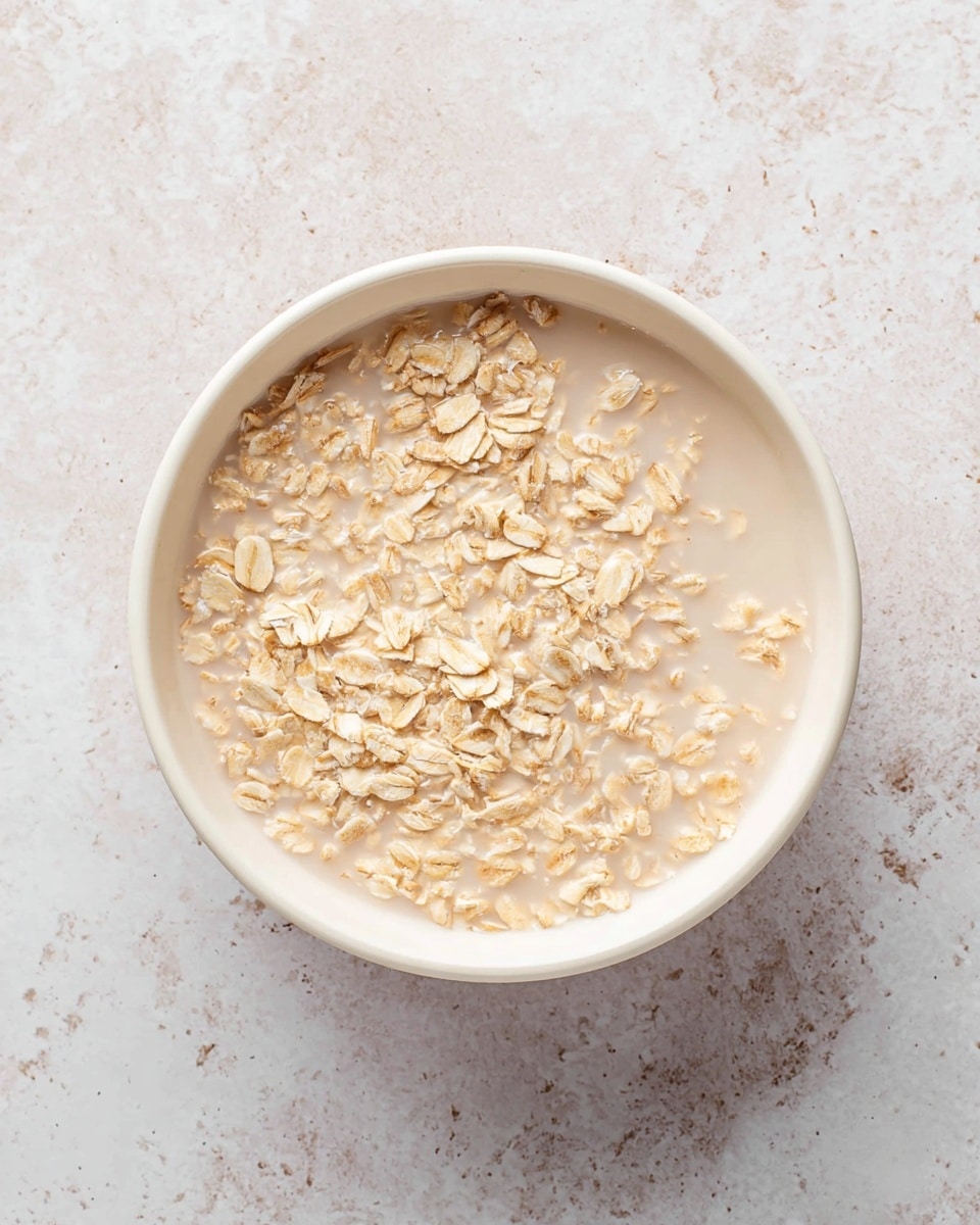 A white bowl is filled with two main layers: the bottom layer is a light beige milk-like liquid, and the top layer is covered with dry beige rolled oats that look rough and spread evenly across the liquid. The bowl is placed on a white marbled textured surface. photo taken with an iphone --ar 4:5 --v 7