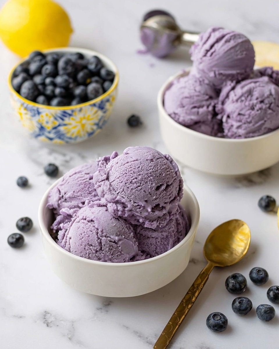 The image shows two white bowls filled with several scoops of light purple ice cream with a textured, creamy surface, placed on a white marbled surface. Behind the bowls, there is a small white bowl with a blue and yellow patterned rim full of dark blue blueberries. A golden spoon with a wooden handle lies beside the front bowl, and there are scattered blueberries around the bowls. Toward the back right, there is a used ice cream scoop with some of the same purple ice cream still inside. A bright yellow lemon sits near the left side of the image, adding a pop of color. The overall scene is bright and inviting, with cool and fresh tones. photo taken with an iphone --ar 4:5 --v 7