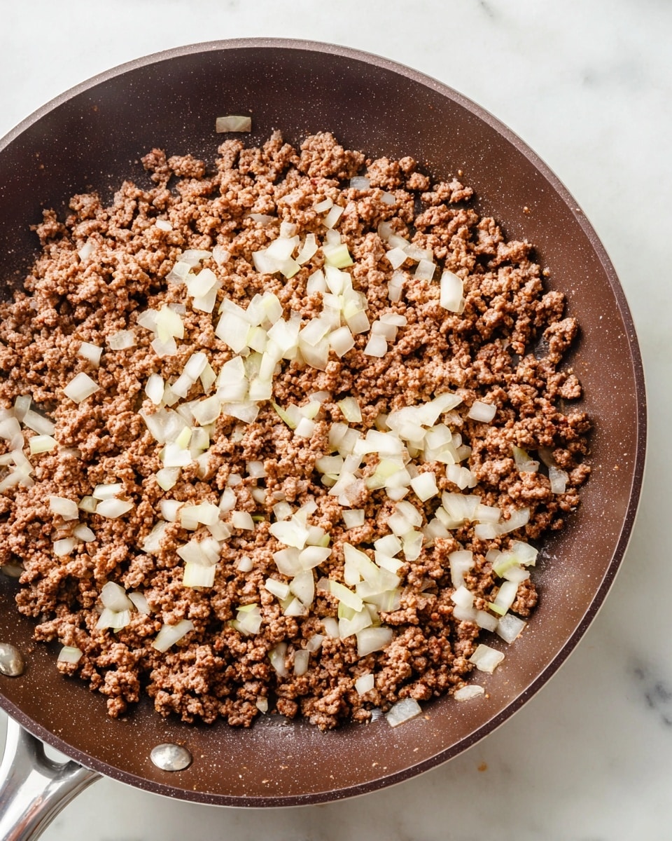 A close-up view of a dark brown frying pan on a white marbled surface, filled with cooked ground meat mixed with small pieces of chopped white onion scattered evenly on top. The meat texture is finely crumbed and lightly browned, while the onions appear soft and translucent, adding contrast with their pale color. The pan handle is partially visible on the left side, extending from the round pan with a slightly shiny finish. Photo taken with an iphone --ar 4:5 --v 7