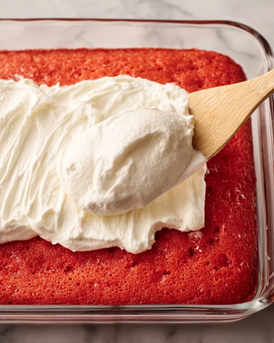 A close-up view of a freshly baked bright red cake in a clear glass baking dish. On top, there is a large dollop of thick, smooth white cream being spread with a wooden spatula. The cake surface shows a soft, slightly textured crumb with a vivid red color, and the creamy layer looks fluffy and glossy as it begins to cover the top. The background has a white marbled texture. photo taken with an iphone --ar 4:5 --v 7